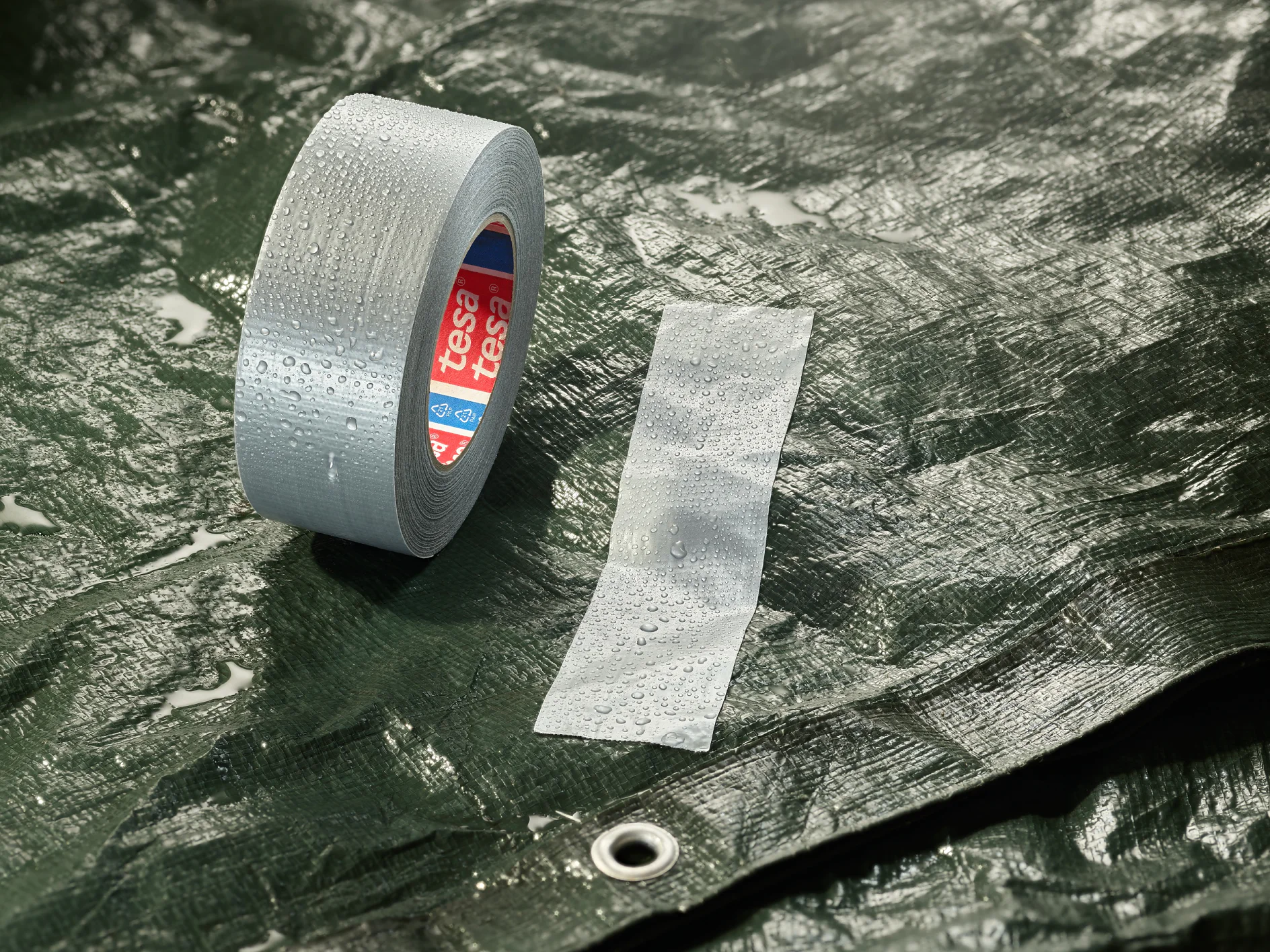 A roll of grey adhesive tesa tape and a strip of tesa tape are placed on a wet, dark green tarpaulin. The tarpaulin has a metal eyelet, and water droplets are visible on the surface. (This text has been generated by AI)