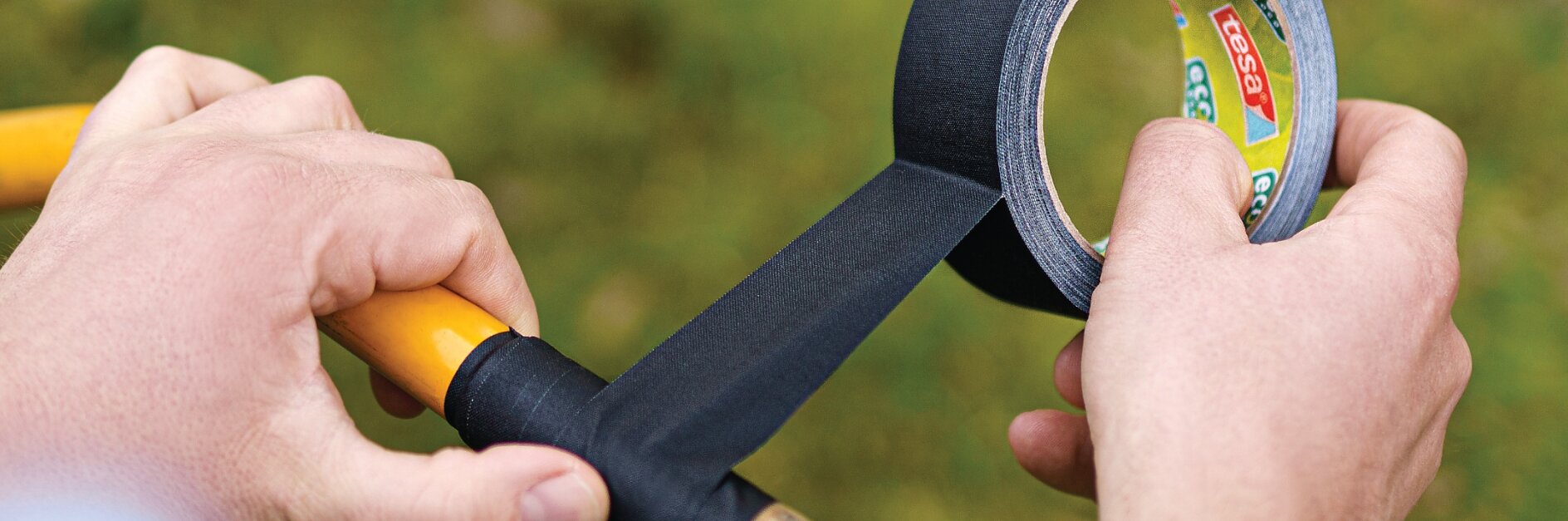 Cilvēks ap dzeltenā instrumenta rokturi aptin melnu lentu. Tesa lente tiek izvilkta no zīmola ruļļa. Fons ir izplūdis ar zaļiem un brūniem toņiem, kas liecina par āra vidi. (This text has been generated by AI)