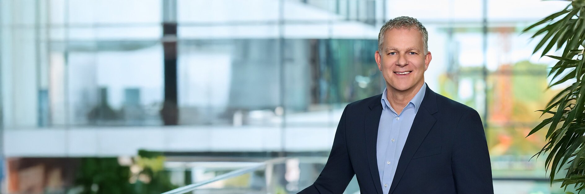 Vīrietis tumšā uzvalkā un gaiši zilā kreklā stāv pie stikla margām iekštelpās, aiz viņa stāv moderna biroju ēka un zaļumi. (This text has been generated by AI)