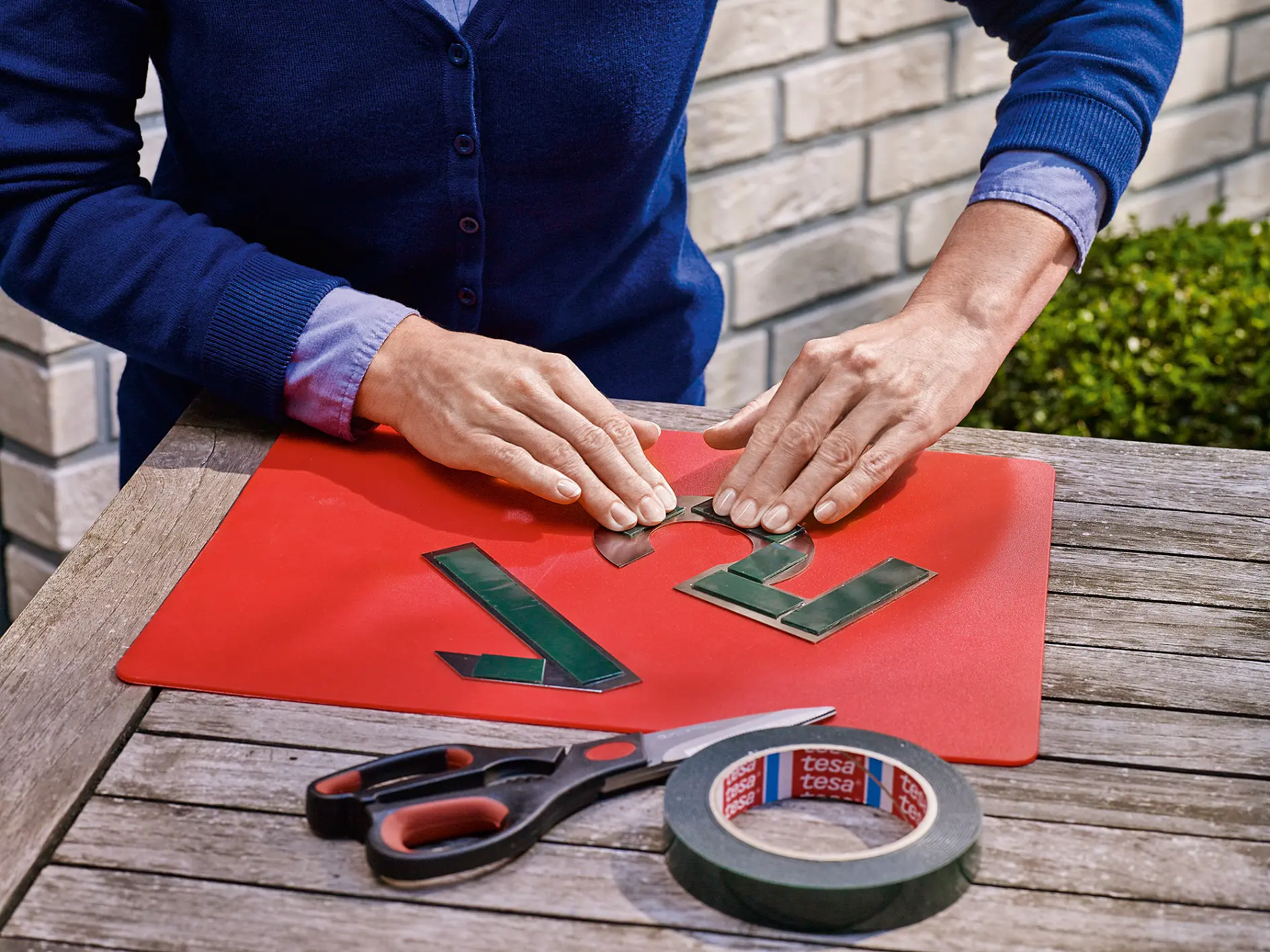 A person in a blue sweater is arranging green adhesive numbers on a red mat on a wooden table. Nearby are scissors, a roll of tesa tape, and a segment of a shrub. Part of a brick wall is visible in the background. (This text has been generated by AI)