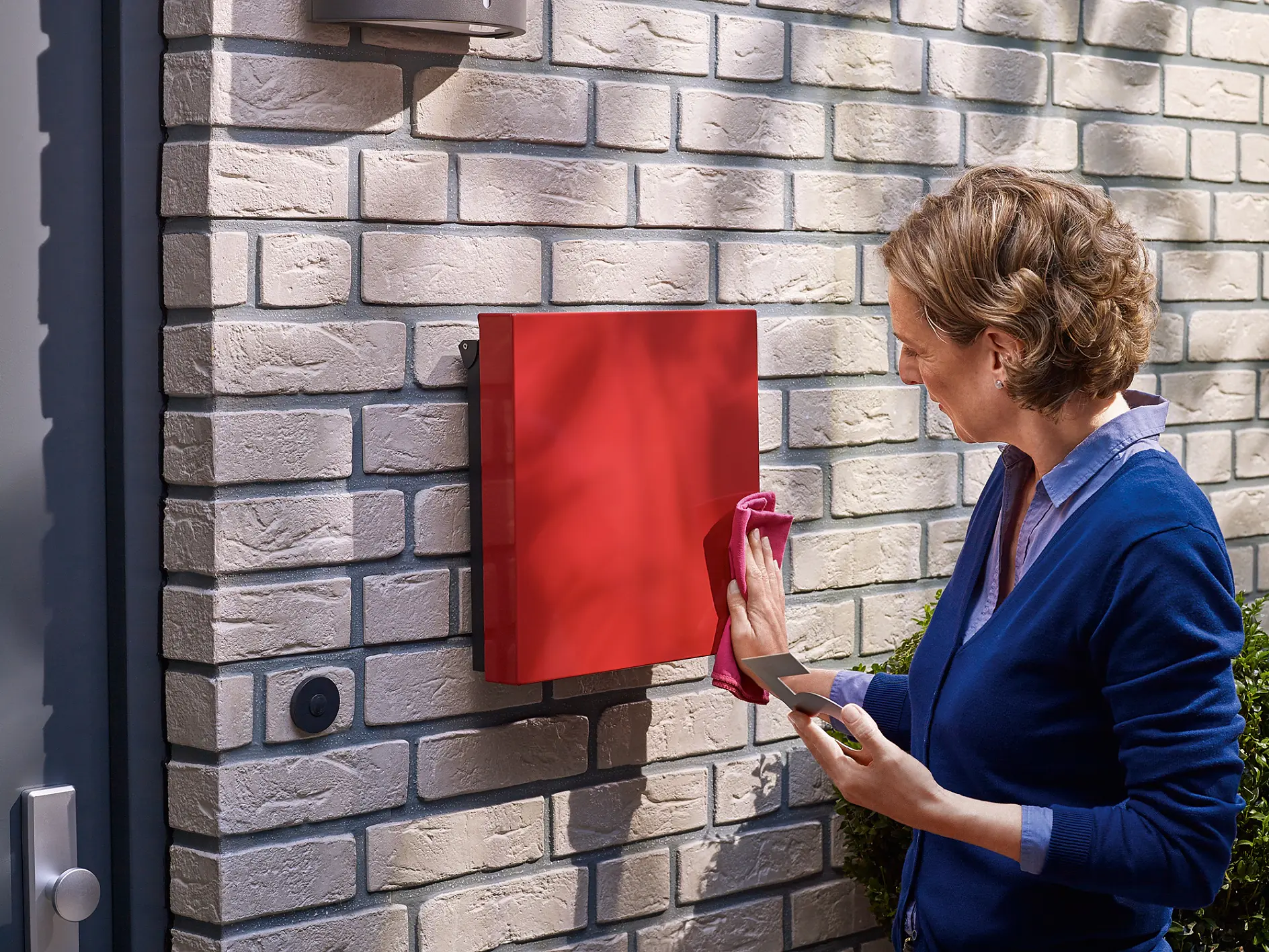 A person in a blue cardigan is cleaning a red mailbox attached to a brick wall. They are using a cloth in one hand while holding tesa tape in the other. A doorbell is visible on the wall next to the mailbox. (This text has been generated by AI)