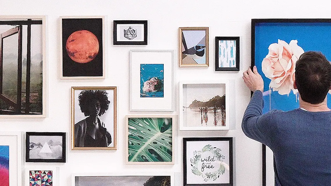 A person in a blue sweater is adjusting a framed picture of a large rose on a blue background on a gallery wall using tesa tape. The wall features various framed artworks, including a red planet, landscapes, abstract designs, and nature images.