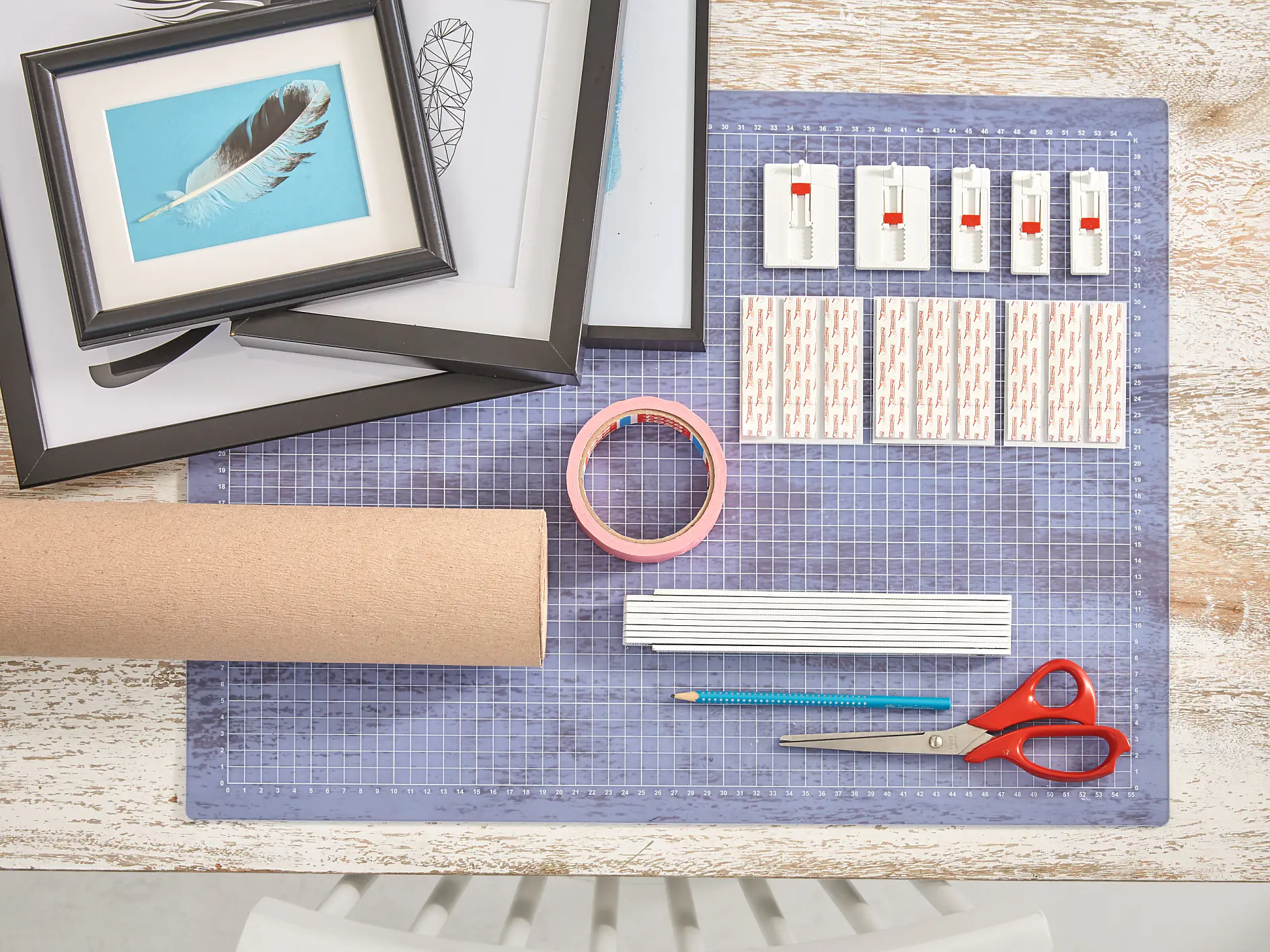 A table with a grid cutting mat, framed pictures, adhesive strips, a cardboard tube, pink tesa tape, a pencil, a ruler, and red-handled scissors. The scene suggests a crafting or DIY project setup. (This text has been generated by AI)