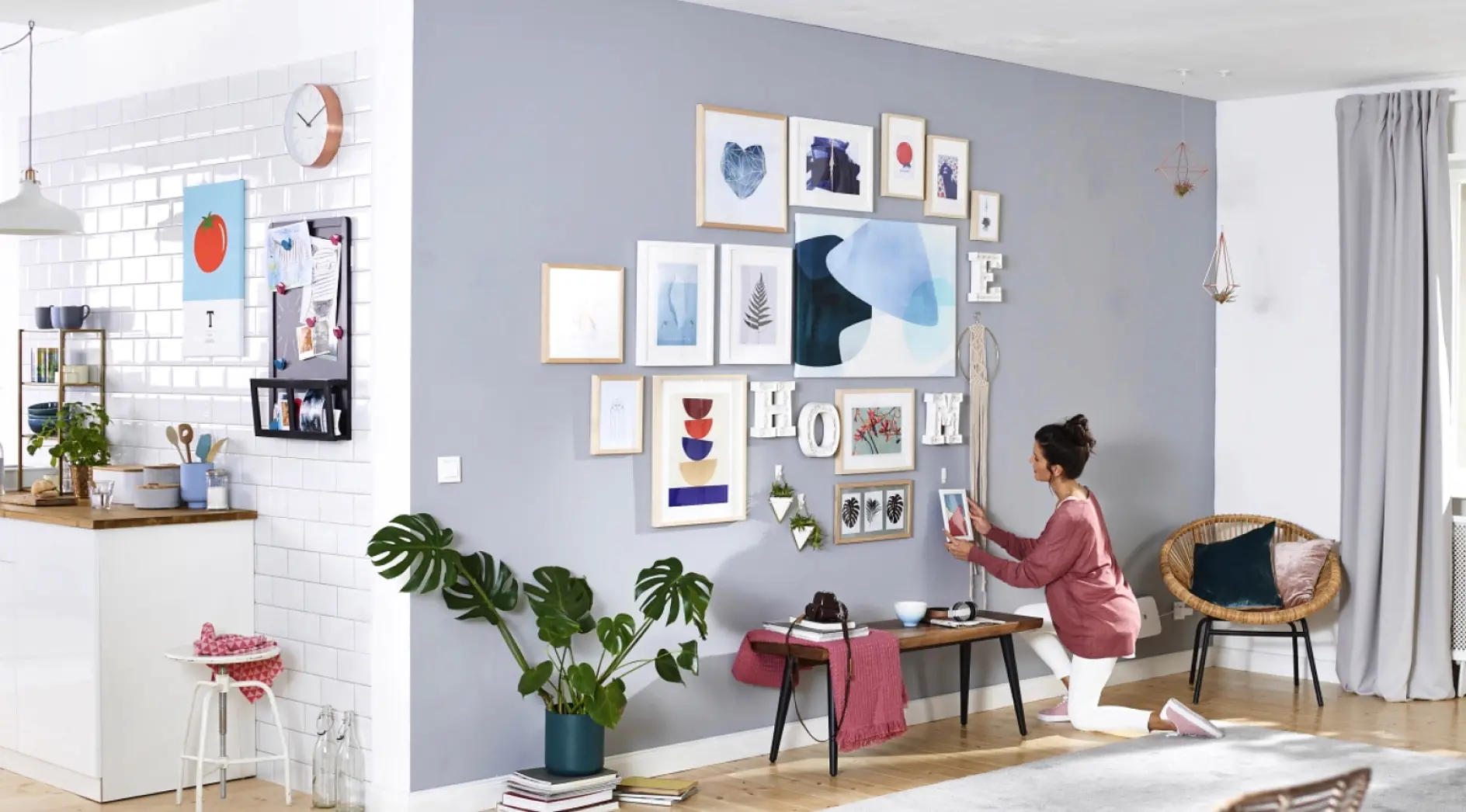 A person kneels on the floor, arranging picture frames on a light gray wall in a modern living area using tesa tape. The room features a plant, a small bench, wicker chair, and a kitchen area in the background. Light wooden flooring and gray curtains are visible. (This text has been generated by AI)
