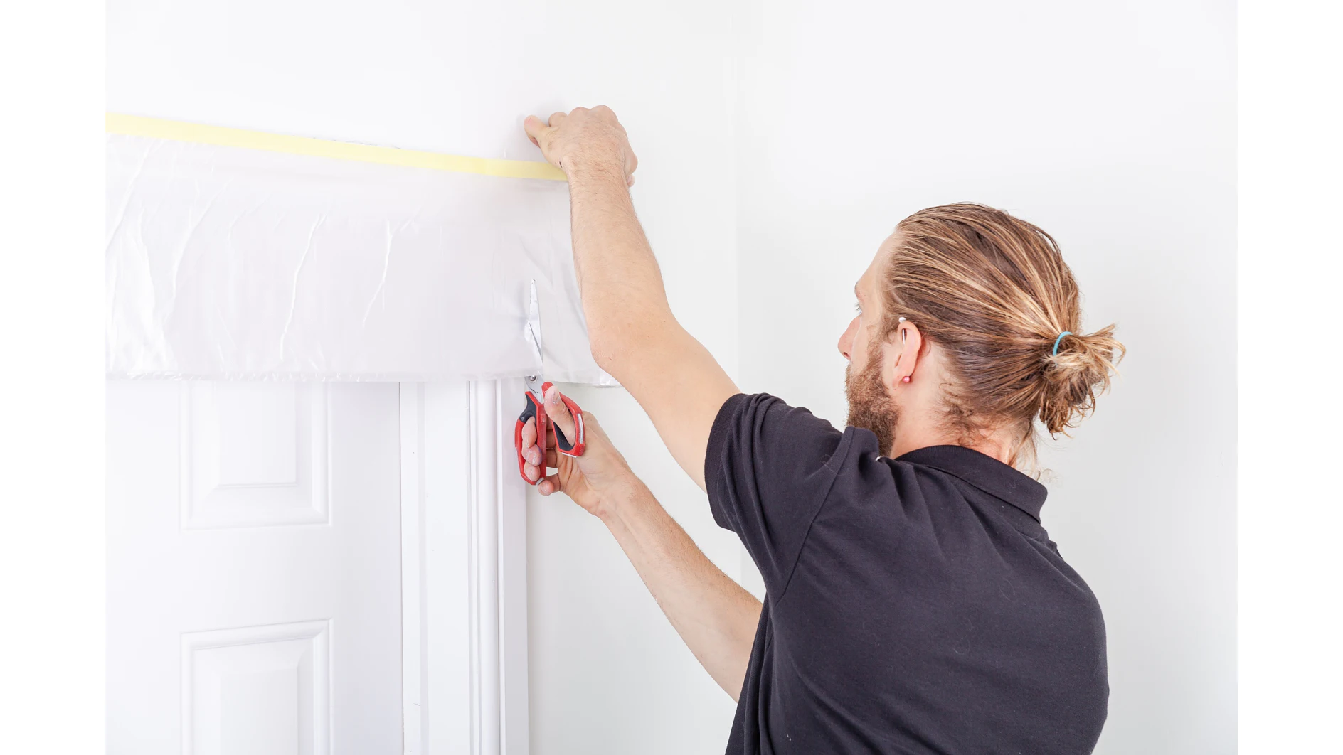 A man with a ponytail is standing by a white door, measuring and cutting a sheet of plastic secured above the doorway with tesa tape. He is using a pair of scissors and wearing a black shirt. The walls and door are white. (This text has been generated by AI)