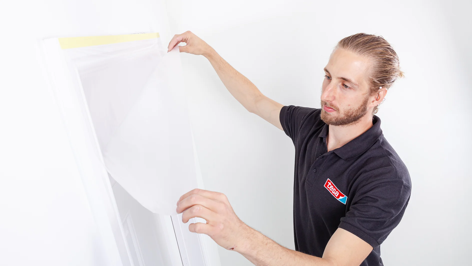 A person with a beard and long hair pulled back, wearing a black polo shirt with a logo, applies a protective sheet over a door frame using tesa tape. The wall and door are white, and the sheet appears to be translucent plastic. (This text has been generated by AI)