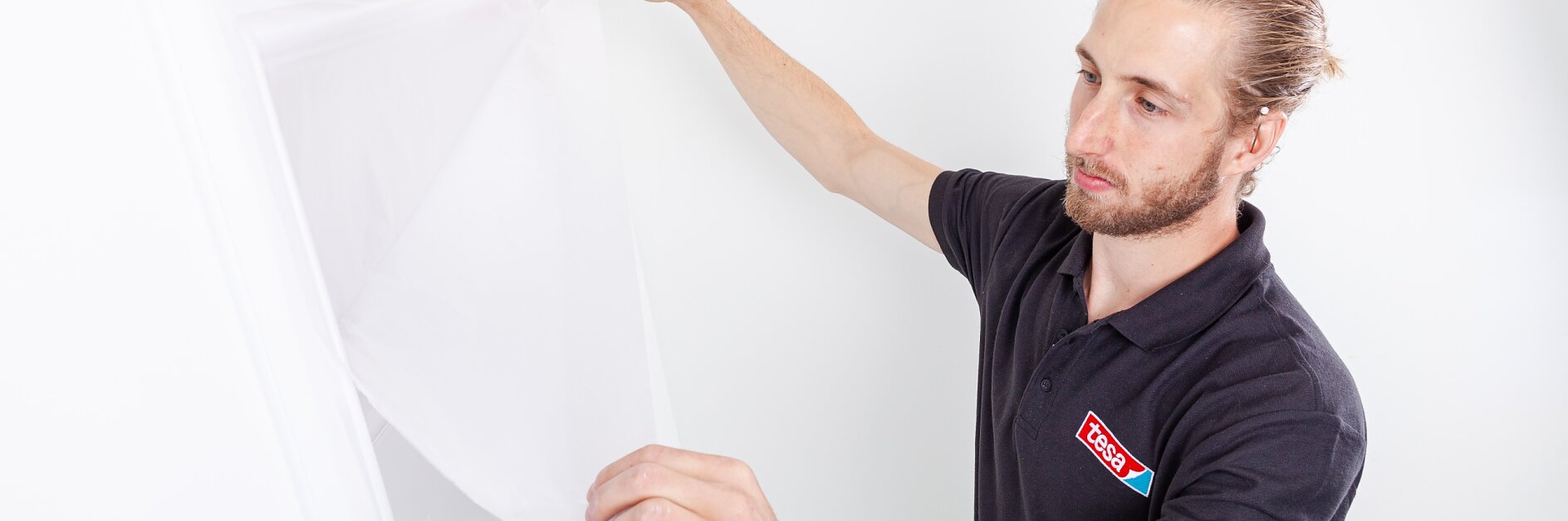 A person with a beard and long hair pulled back, wearing a black polo shirt with a logo, applies a protective sheet over a door frame using tesa tape. The wall and door are white, and the sheet appears to be translucent plastic. (This text has been generated by AI)