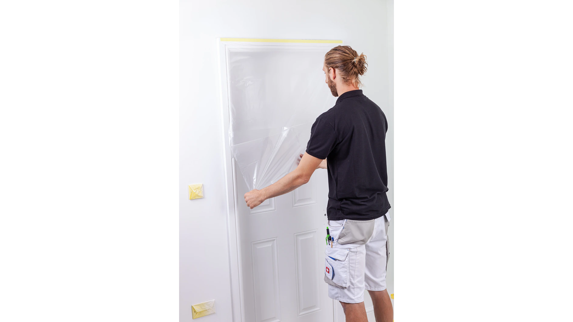 A person in a black shirt and white shorts is removing a plastic sheet from a white door. The wall is light-colored and a light switch is partially covered with tesa tape. Various tools are in the persons pocket. (This text has been generated by AI)