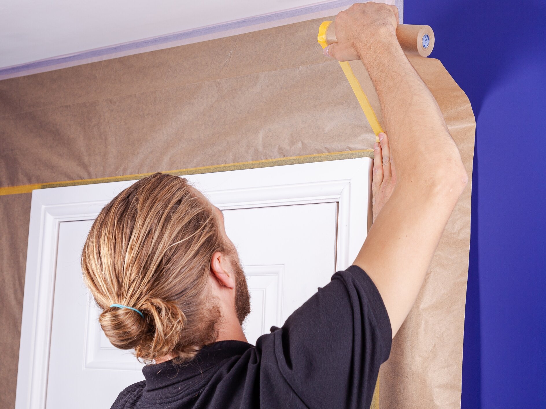A person with long hair in a bun is applying tesa tape to a brown paper covering around a door frame. The wall is painted blue. They are wearing a black shirt and holding the tesa tape in their right hand. (This text has been generated by AI)