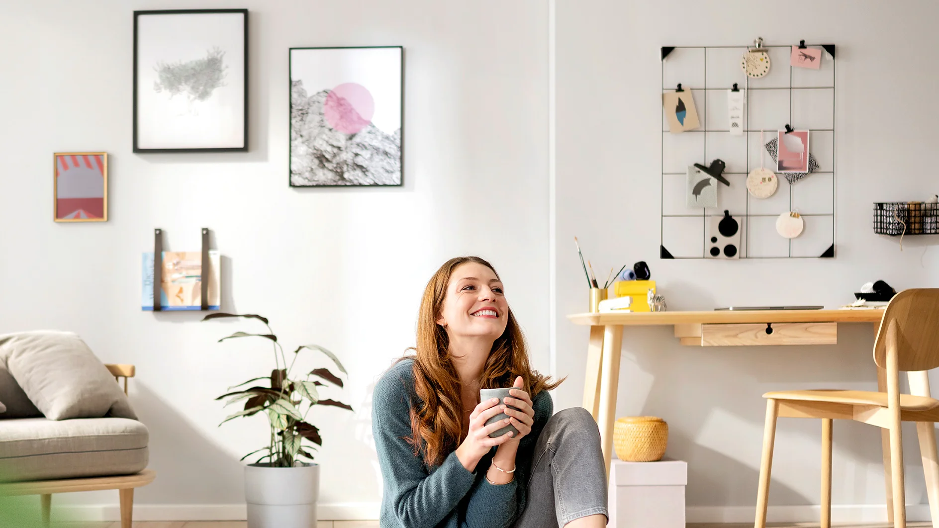 A woman with long hair is sitting on the floor in a brightly lit living room holding a mug, smiling upward. The room features a small desk with a chair, a potted plant, wall art, and decorative items secured with tesa tape on a wire grid. (This text has been generated by AI)