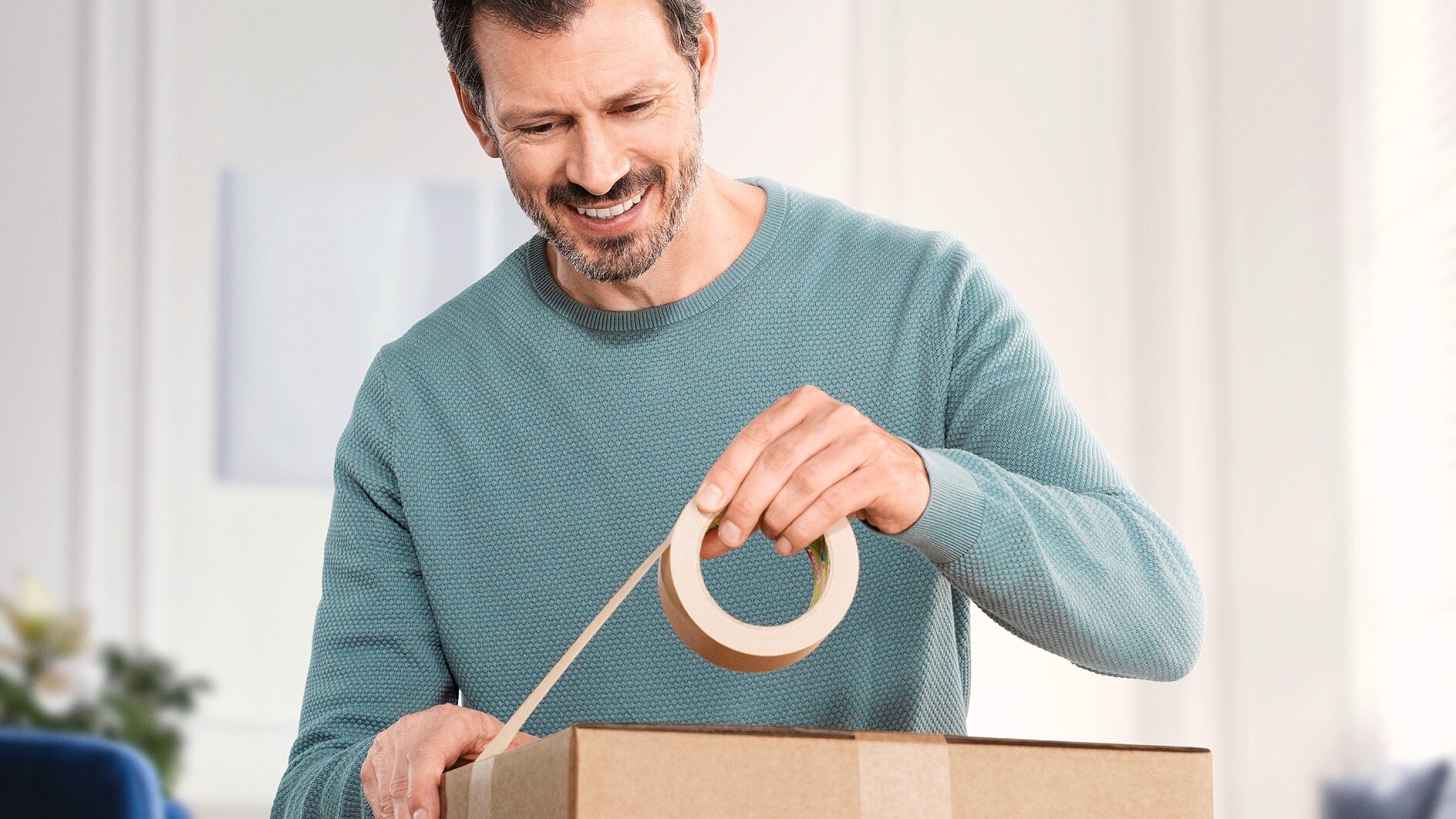 A man in a teal sweater is sealing a cardboard box with tesa tape. He is smiling and appears to be indoors with a blurred background. (This text has been generated by AI)