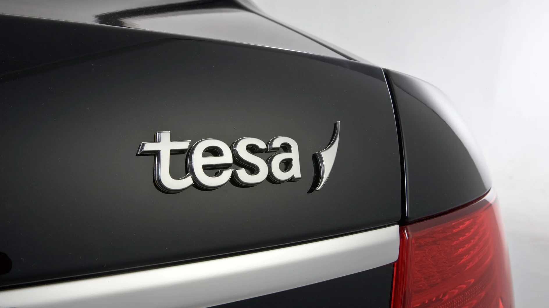 Close-up of the rear end of a black vehicle showing the brand logo tesa tape on the trunk. The design features chrome lettering, and the right tail light is partially visible. The background is plain and neutral. (This text has been generated by AI)