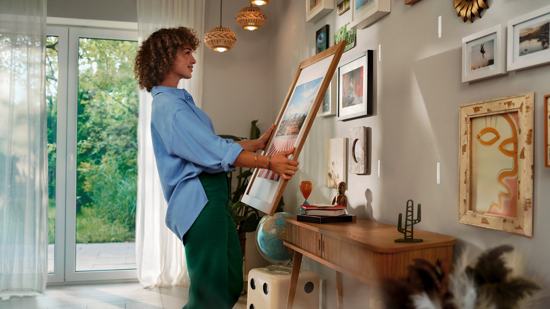 A person in a blue shirt and green pants is hanging a framed picture on a wall using tesa tape. The room is decorated with multiple artworks and has a wooden desk with various items. A window with sheer curtains is open to a view of greenery outside. (This text has been generated by AI)