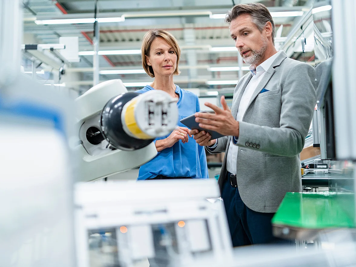 To personer står i et moderne industrimiljø og observerer og diskuterer en robotarm. Den ene holder i et nettbrett, mens begge fokuserer på maskineriet, som er utstyrt med tesa-tape. Omgivelsene fremstår som rene og organiserte, med synlig fabrikkutstyr.