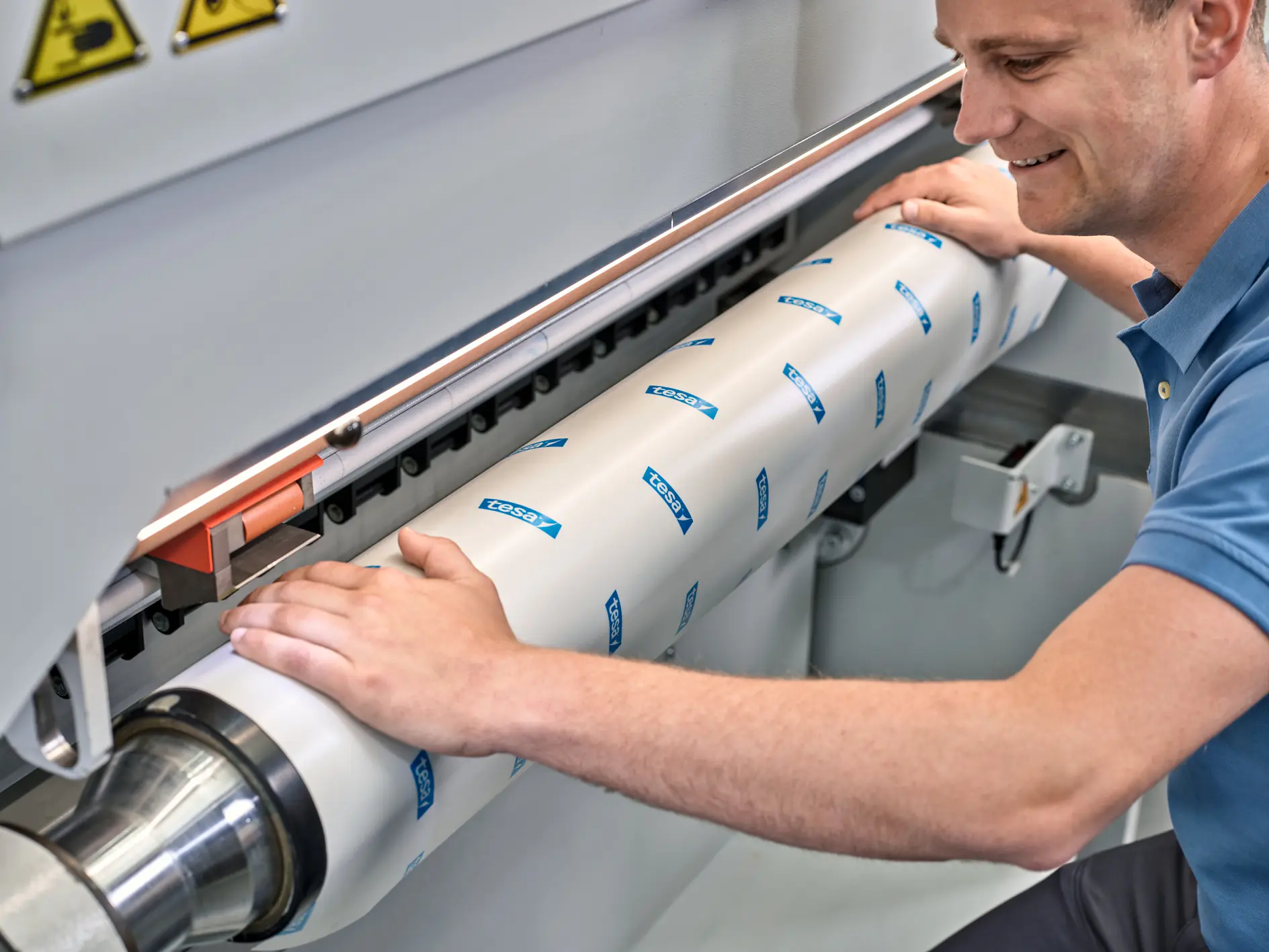 En person betjener en maskin og justerer en stor sylindrisk komponent dekket med hvitt materiale og blå tesa-logoer. Komponenten er festet på plass. Personen er fokusert på oppgaven og har på seg en lyseblå kortermet skjorte. (This text has been generated by AI)
