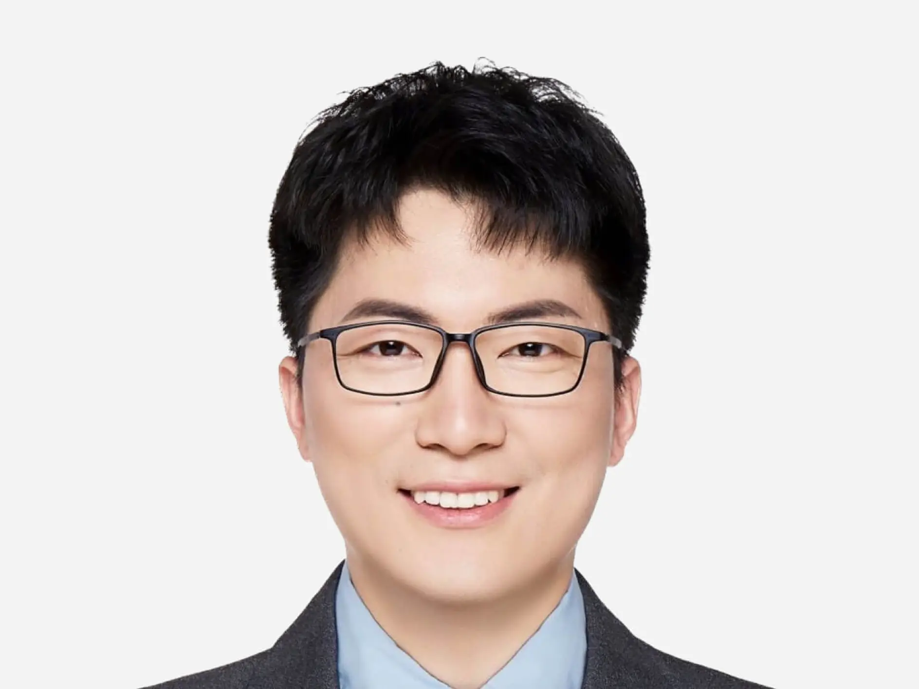 A person with short black hair and glasses is smiling. They are wearing a gray suit and a light blue shirt. The background is plain white, and theyre holding a roll of tesa tape in their hand, showcasing its versatility for various applications.
