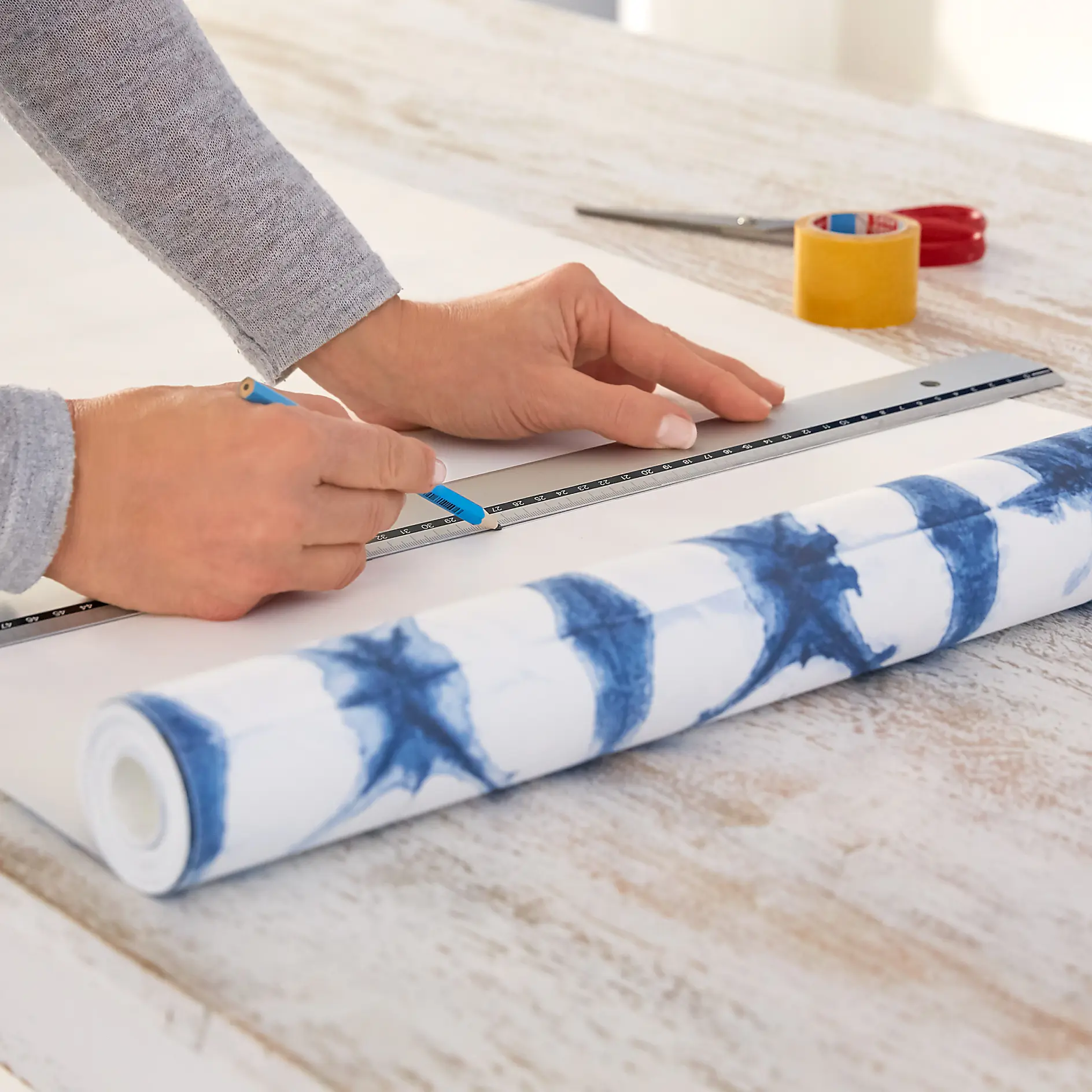 Hands measuring and marking wallpaper with a ruler and pencil on a wooden table. A roll of blue and white patterned wallpaper, scissors, and tesa tape are visible. (This text has been generated by AI)
