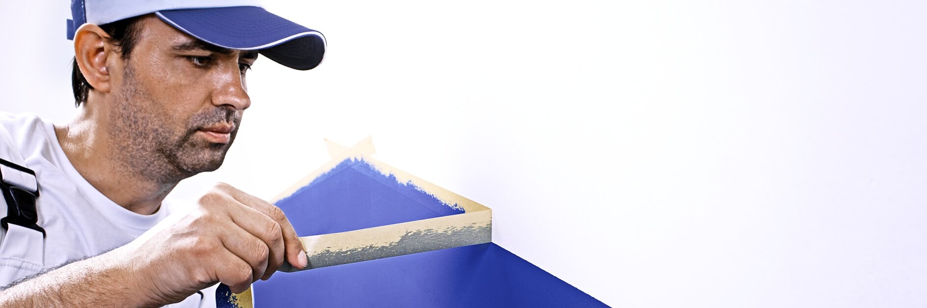 A man wearing a blue cap and a white shirt with blue overalls is painting a geometric design on a wall. He is peeling tesa tape from a blue-painted section on a white wall. A paint can is visible at his side.