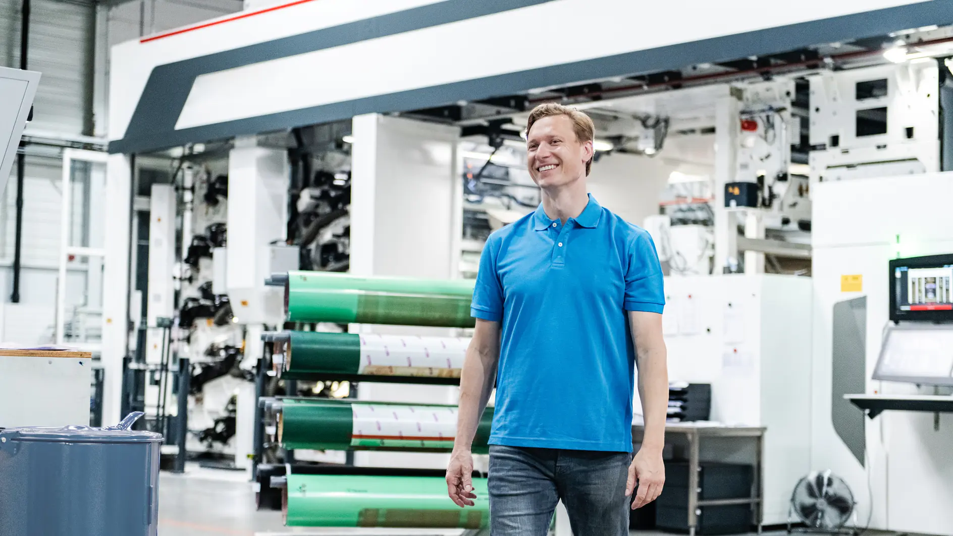 A man in a blue polo shirt and jeans is standing in a large industrial facility. Behind him, there is machinery and large rolls of tesa tape material. The setting appears to be a printing or manufacturing plant.