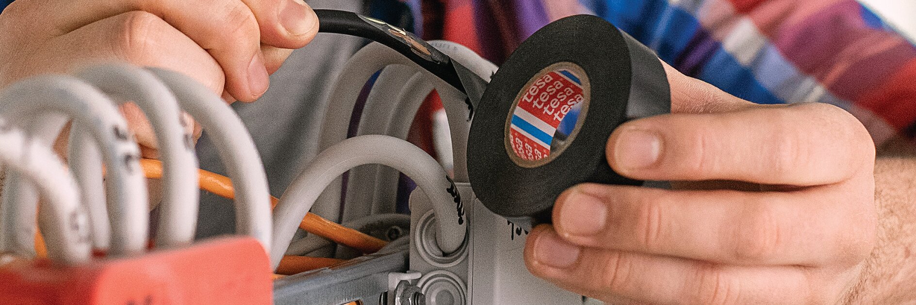 A person wearing a plaid shirt is holding a roll of black tesa tape and is applying it to wires in an electrical box. The scene appears to involve electrical work or installation, with multiple cables being organized.