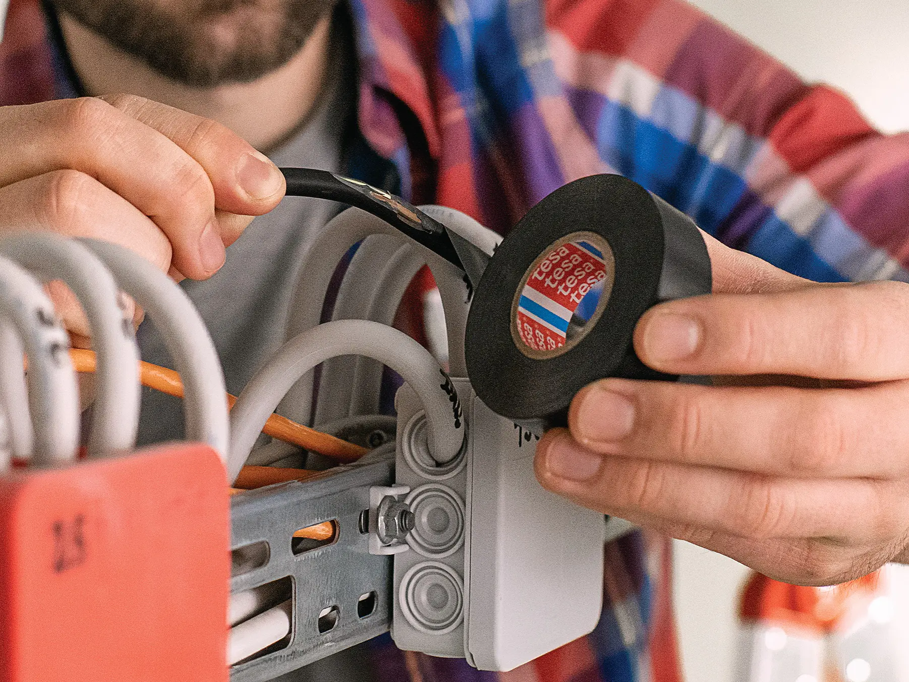 A person wearing a plaid shirt is holding a roll of black tesa tape and is applying it to wires in an electrical box. The scene appears to involve electrical work or installation, with multiple cables being organized.