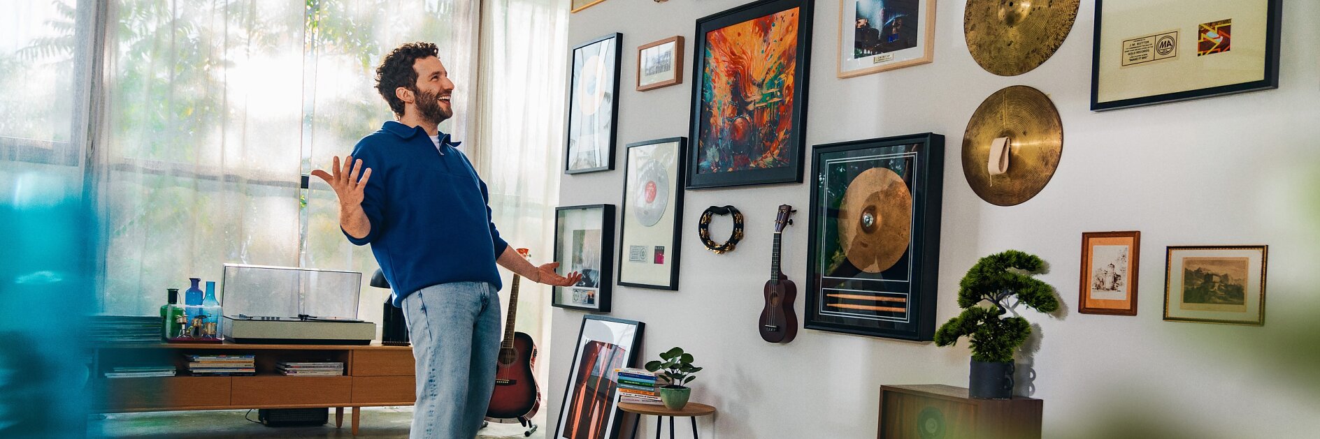 Een persoon staat in een kamer met een speelse pose, omringd door een verscheidenheid aan muurkunst, ingelijste platen en muziekinstrumenten. Een gitaar rust tegen de muur en er zijn speakers en een draaitafel op een plank. De kamer is goed verlicht door natuurlijk zonlicht. (This text has been generated by AI)