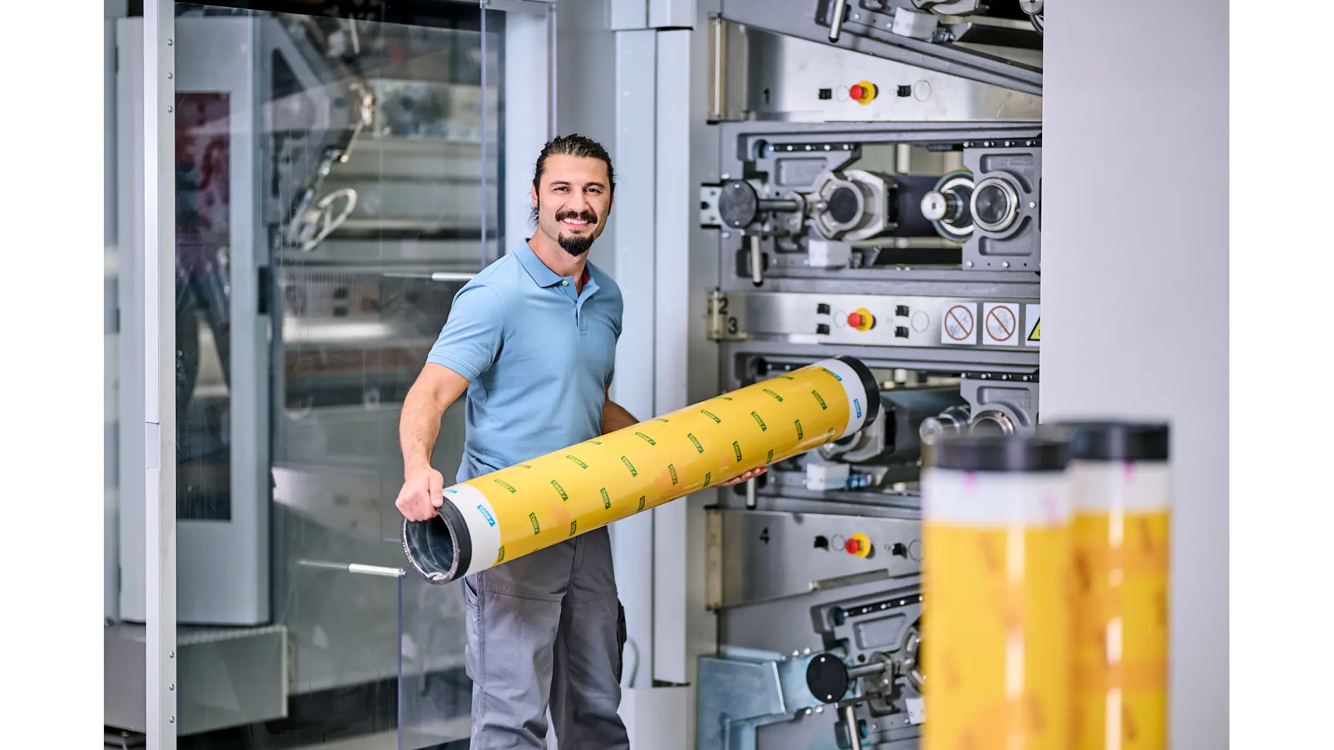 A man with a beard is holding a large cylindrical roll of tesa tape with yellow and black markings. He stands next to industrial printing machinery inside a manufacturing facility. He is wearing a light blue shirt and gray pants. (This text has been generated by AI)