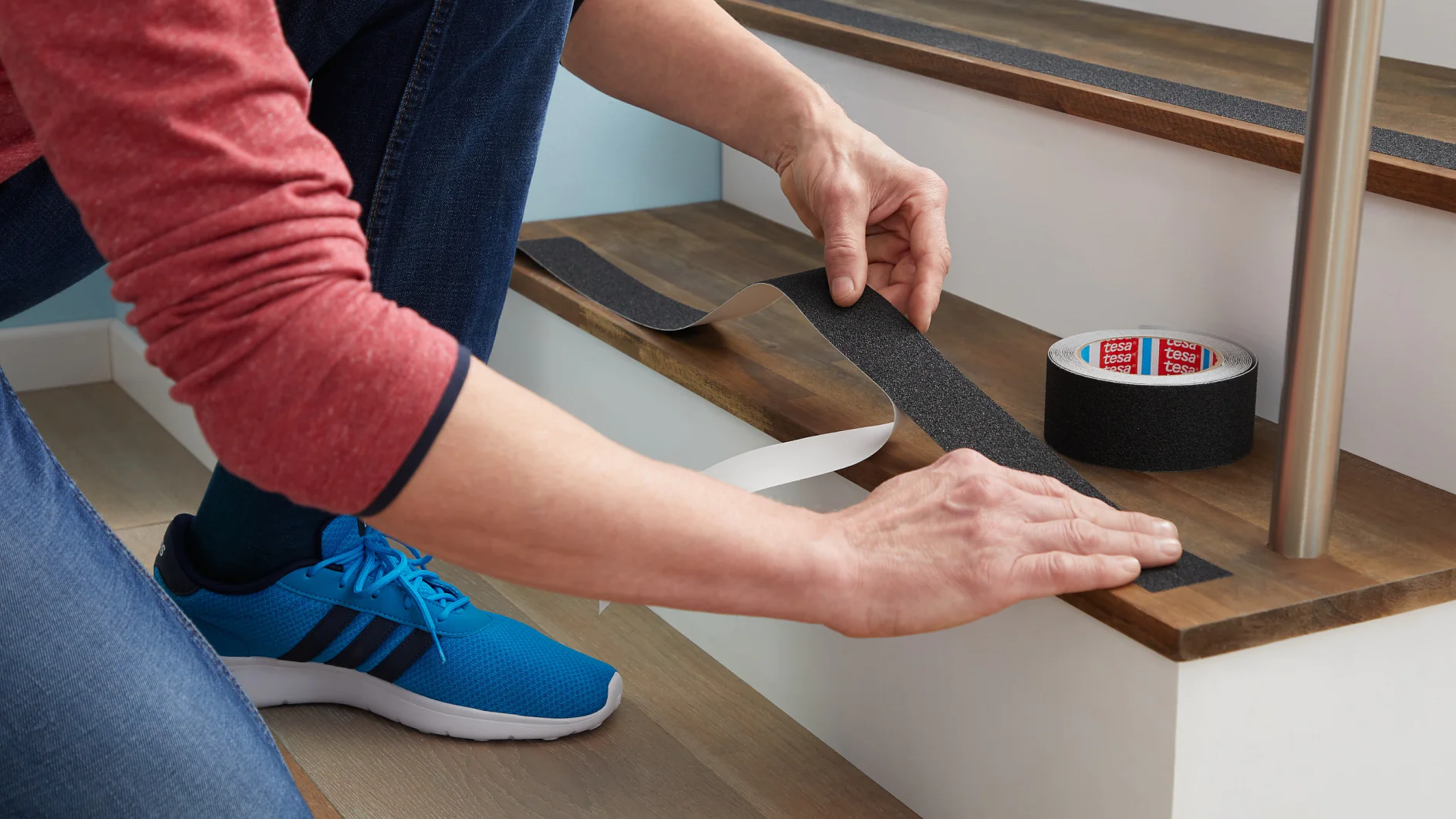 Een persoon plakt een strook zwarte antislip tesa-tape op de rand van een houten traptrede, met een rol tesa-tape en een gedeeltelijk verwijderde beschermlaag zichtbaar. De persoon draagt blauwe sneakers en een spijkerbroek. (This text has been generated by AI)