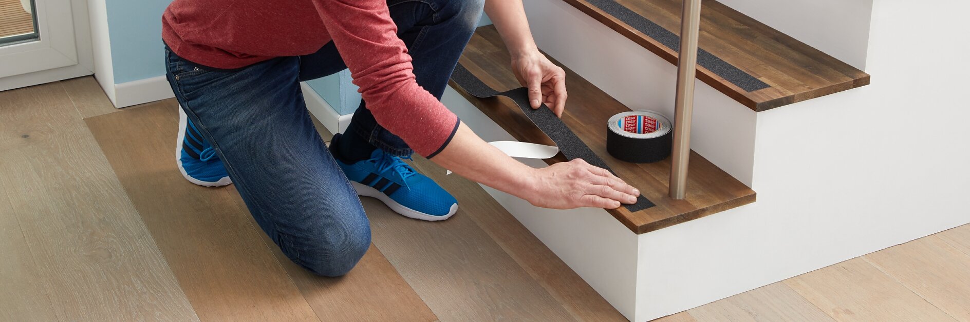 Een man in een rood overhemd en een blauwe spijkerbroek knielt op de grond naast een trap en plakt zwarte tesa-tape op de rand van een houten traptrede. Rollen tesa-tape liggen op de trede naast hem. (This text has been generated by AI)