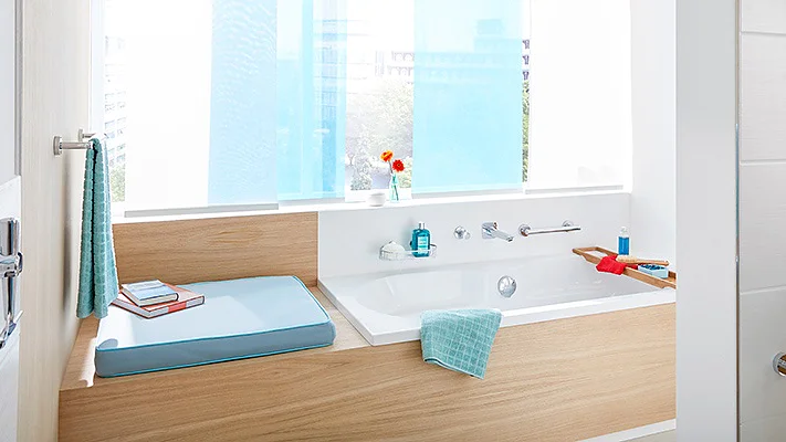 A modern bathroom features a wooden-accented bathtub next to a window with blue curtains. A bath tray holds a red book and blue towel. Toiletries and a potted plant are arranged neatly on the shelf. The space is bright and well-organized, with tesa tape ensuring seamless finishes throughout the room. (This text has been generated by AI)