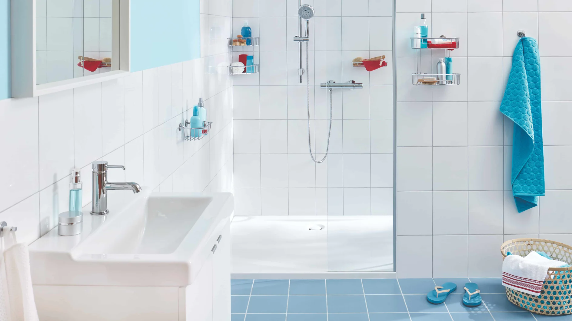 A modern bathroom features white tile walls and a white sink. The shower area includes a glass divider, a handheld showerhead, and shelves for toiletries. A blue towel is neatly hung on the right, accompanied by matching slippers and a laundry basket on the blue-tiled floor. (This text has been generated by AI)