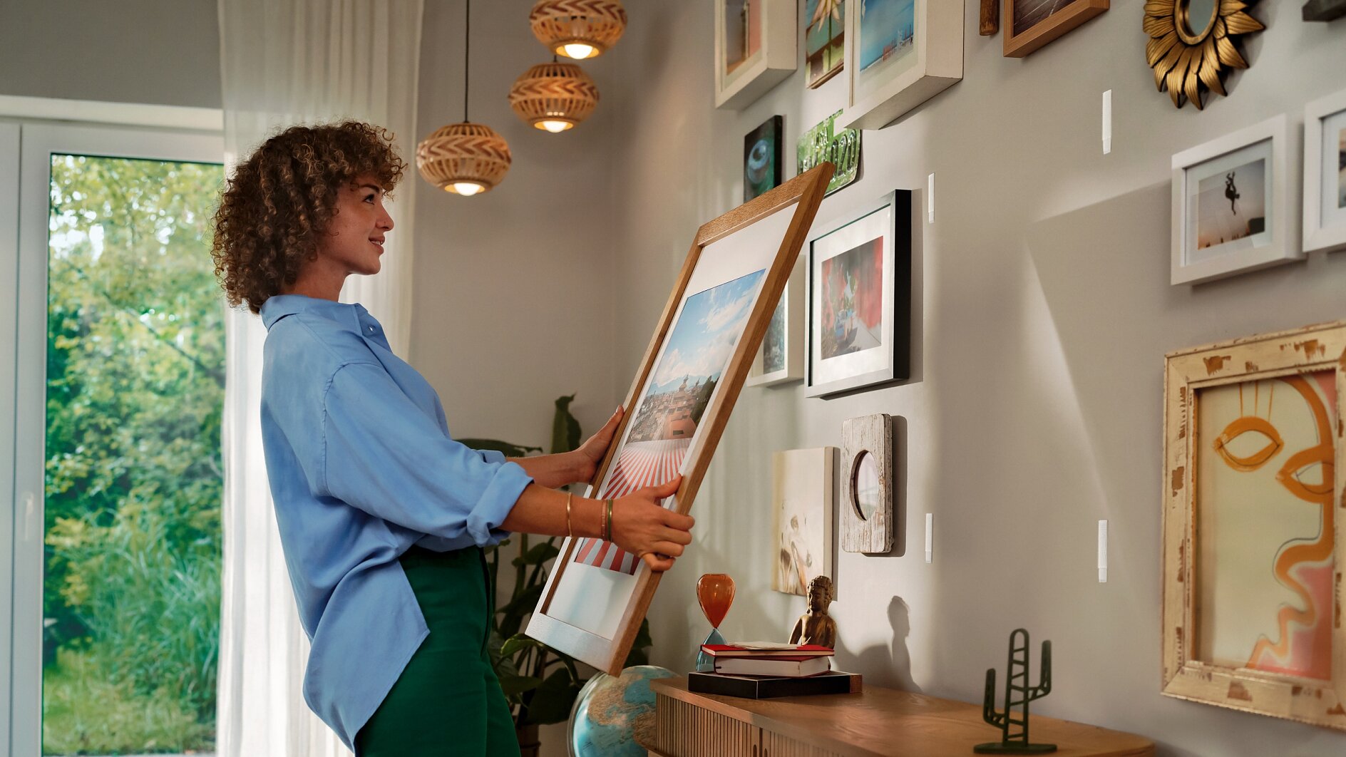 Een vrouw met krullend haar hangt een ingelijste foto op een witte muur versierd met verschillende kunstwerken en foto's, met behulp van tesa tape, naast een houten dressoir met decoratieve voorwerpen, onder warme hanglampen. (This text has been generated by AI)