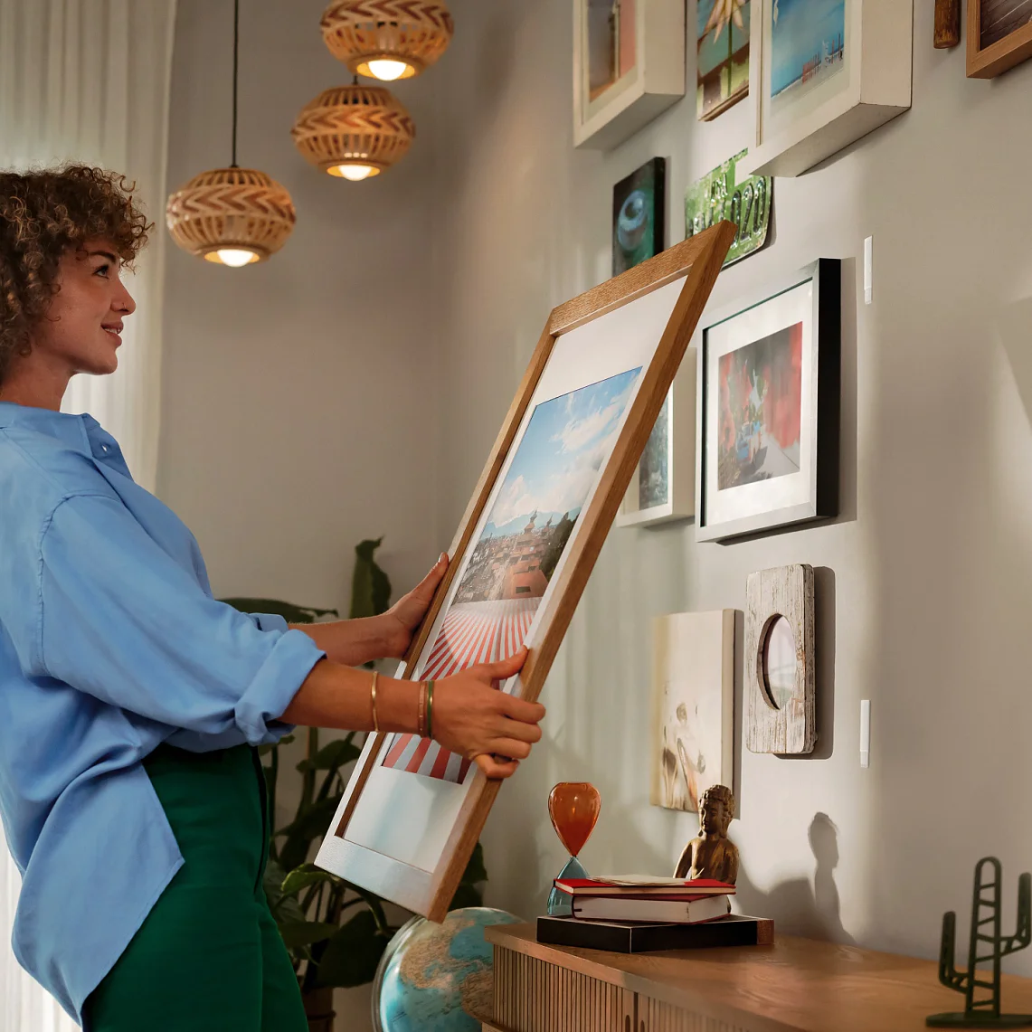 Een vrouw met krullend haar hangt een ingelijste foto op een witte muur versierd met verschillende kunstwerken en foto's, met behulp van tesa tape, naast een houten dressoir met decoratieve voorwerpen, onder warme hanglampen. (This text has been generated by AI)