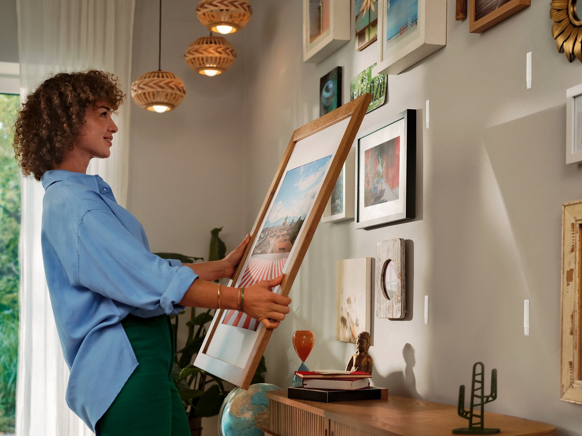 Een vrouw met krullend haar hangt een ingelijste foto op een witte muur versierd met verschillende kunstwerken en foto's, met behulp van tesa tape, naast een houten dressoir met decoratieve voorwerpen, onder warme hanglampen. (This text has been generated by AI)