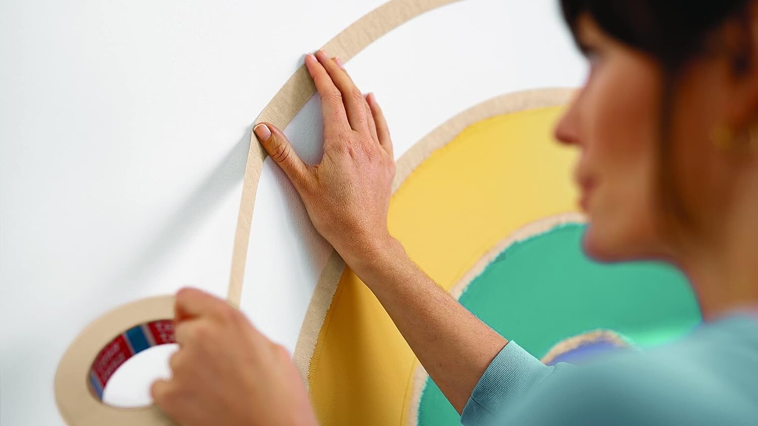 Een persoon drukt tesa tape op een witte muur en omlijnt zo de rand van een veelkleurig cirkelvormig ontwerp met gele, groene en groenblauwe delen. De rol tesa tape wordt in de linkerhand gehouden. (This text has been generated by AI)