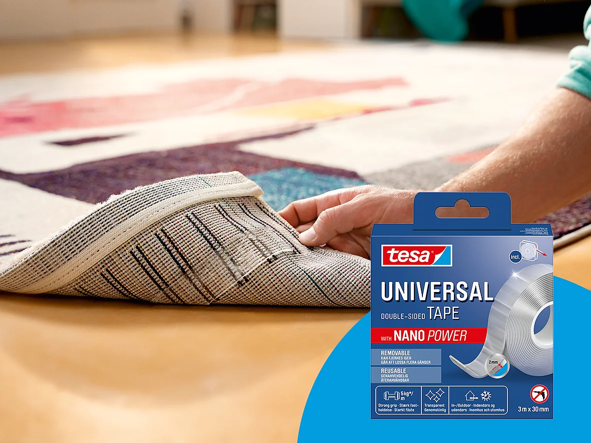 Een hand tilt de hoek van een gedessineerd tapijt op en onthult dubbelzijdige tesa tape waarmee het op een houten vloer wordt vastgezet. Rechtsonder staat een doos tesa Universal Tape met Nano Power. (This text has been generated by AI)