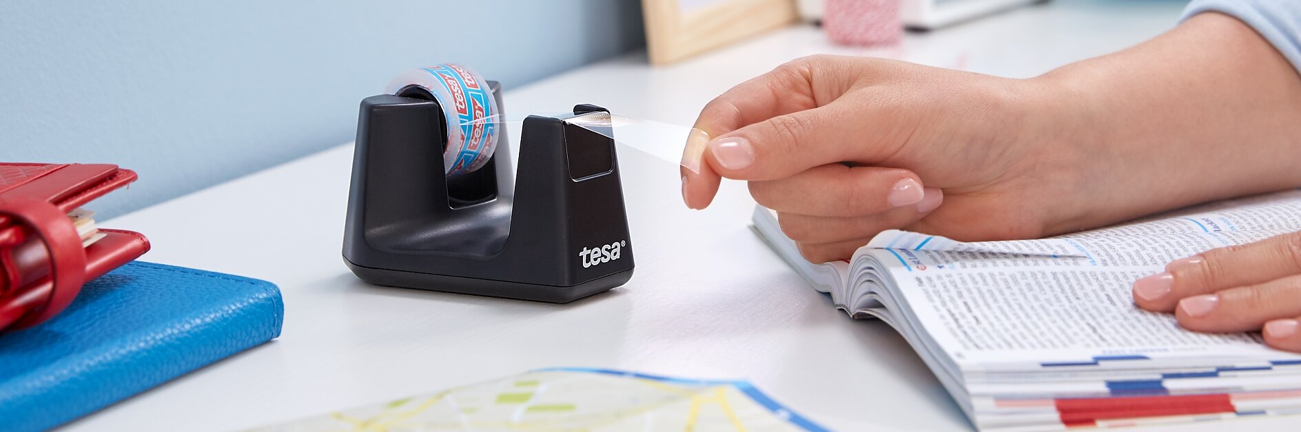 Een persoon trekt een strook decoratieve tesa tape uit een zwarte tesa tape dispenser op een bureau, met een open tijdschrift, een rood notitieblok, een blauwe map en een kaart in de buurt. (This text has been generated by AI)