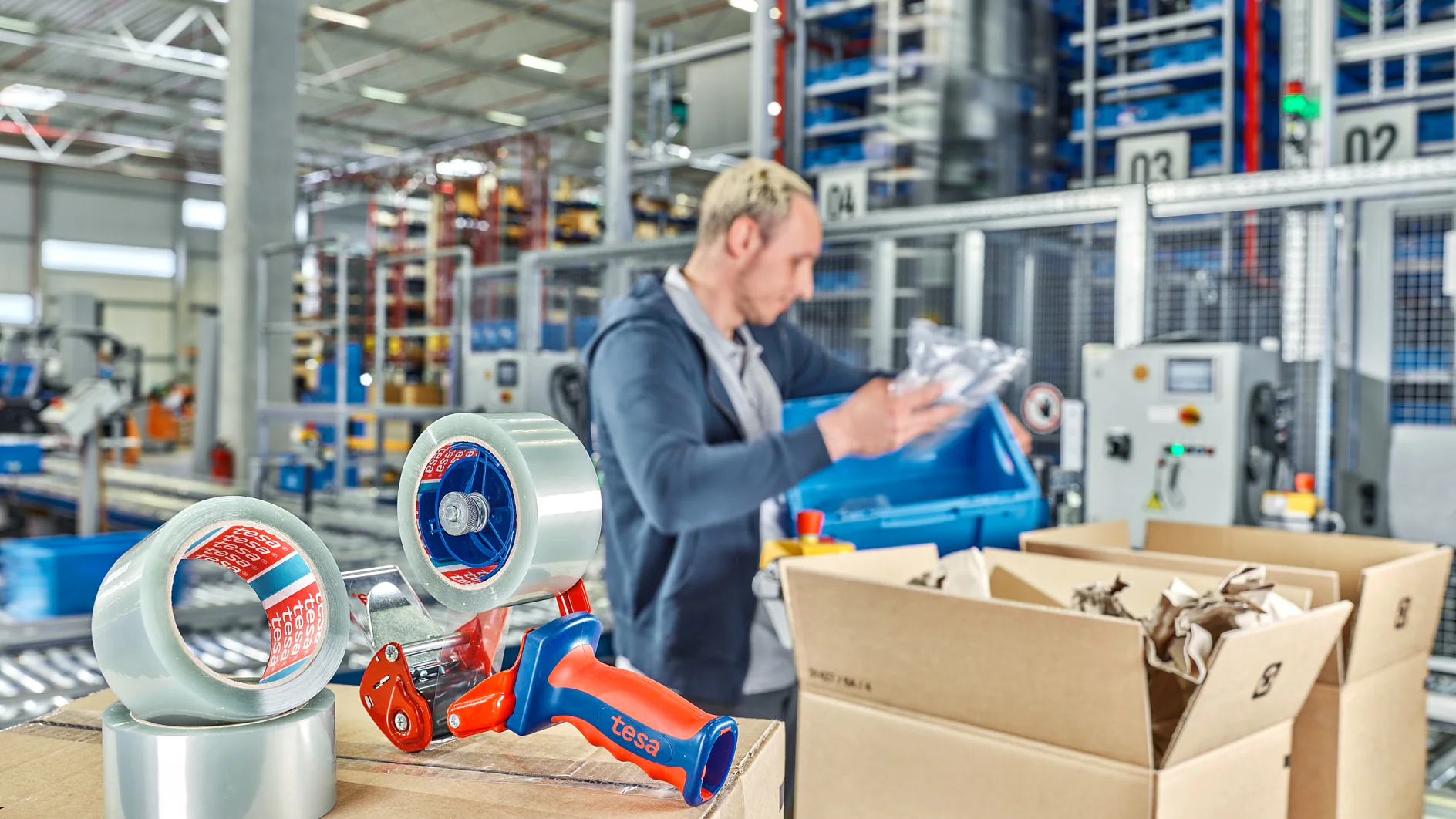 Een persoon in een magazijn staat bij lopende banden dozen in te pakken. Twee rollen tesa tape en een dispenser zitten op een doos vooraan. (This text has been generated by AI)