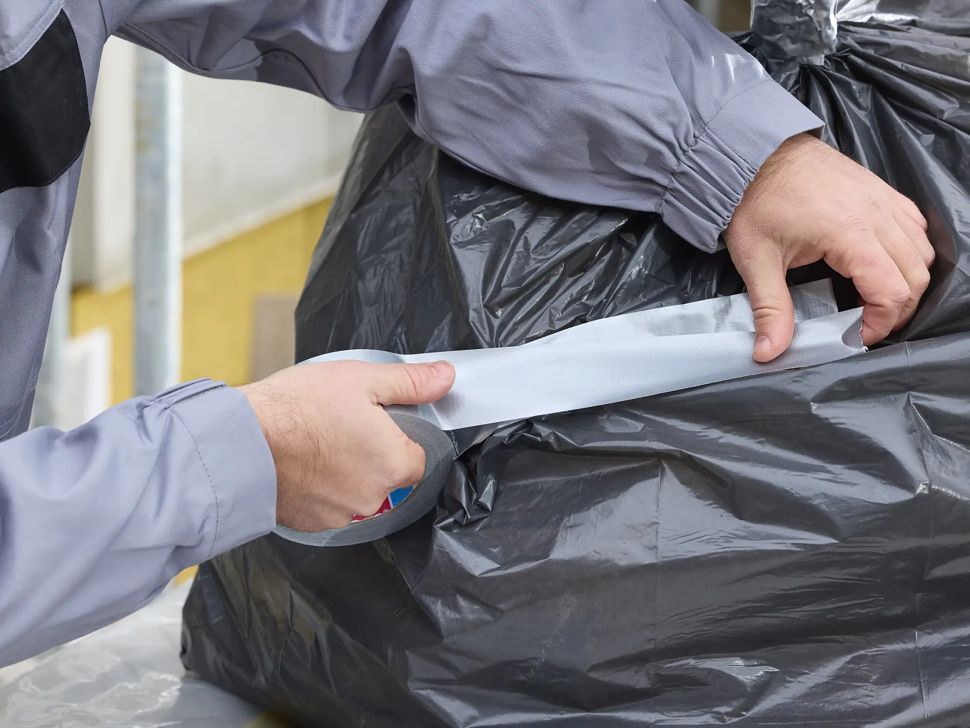 hands in gray sleeves sealing a black trash bag with silver tesa tape in an indoor setting (This text has been generated by AI)