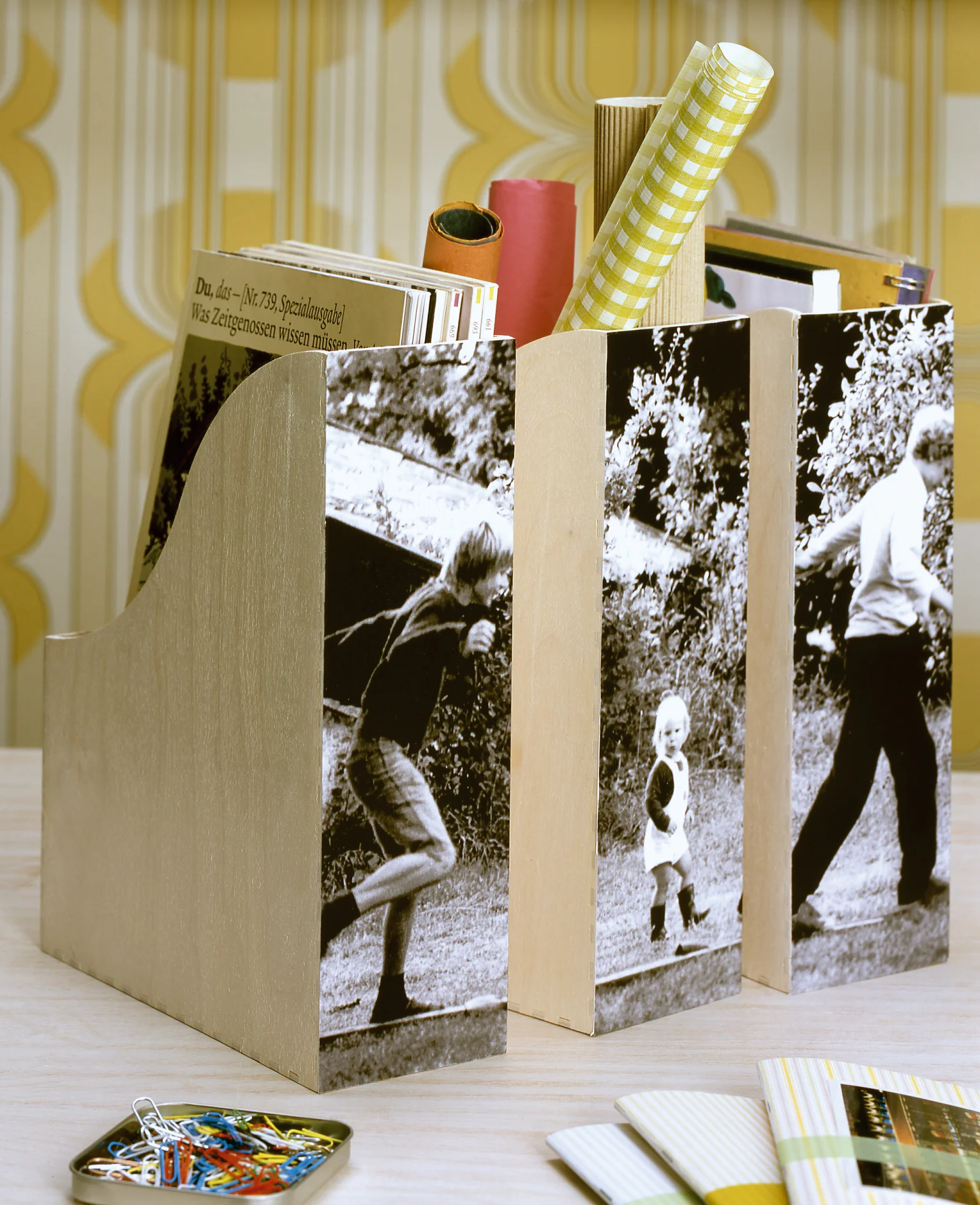 Houten tijdschriftenhouders staan op een bureau, elk versierd met zwart-witfotos van mensen die buiten lopen. Ze bevatten verschillende items, waaronder opgerold papier vastgehouden met tesa-tape en boeken. Een set sleutels en kleurstalen liggen op het bureau. (This text has been generated by AI)