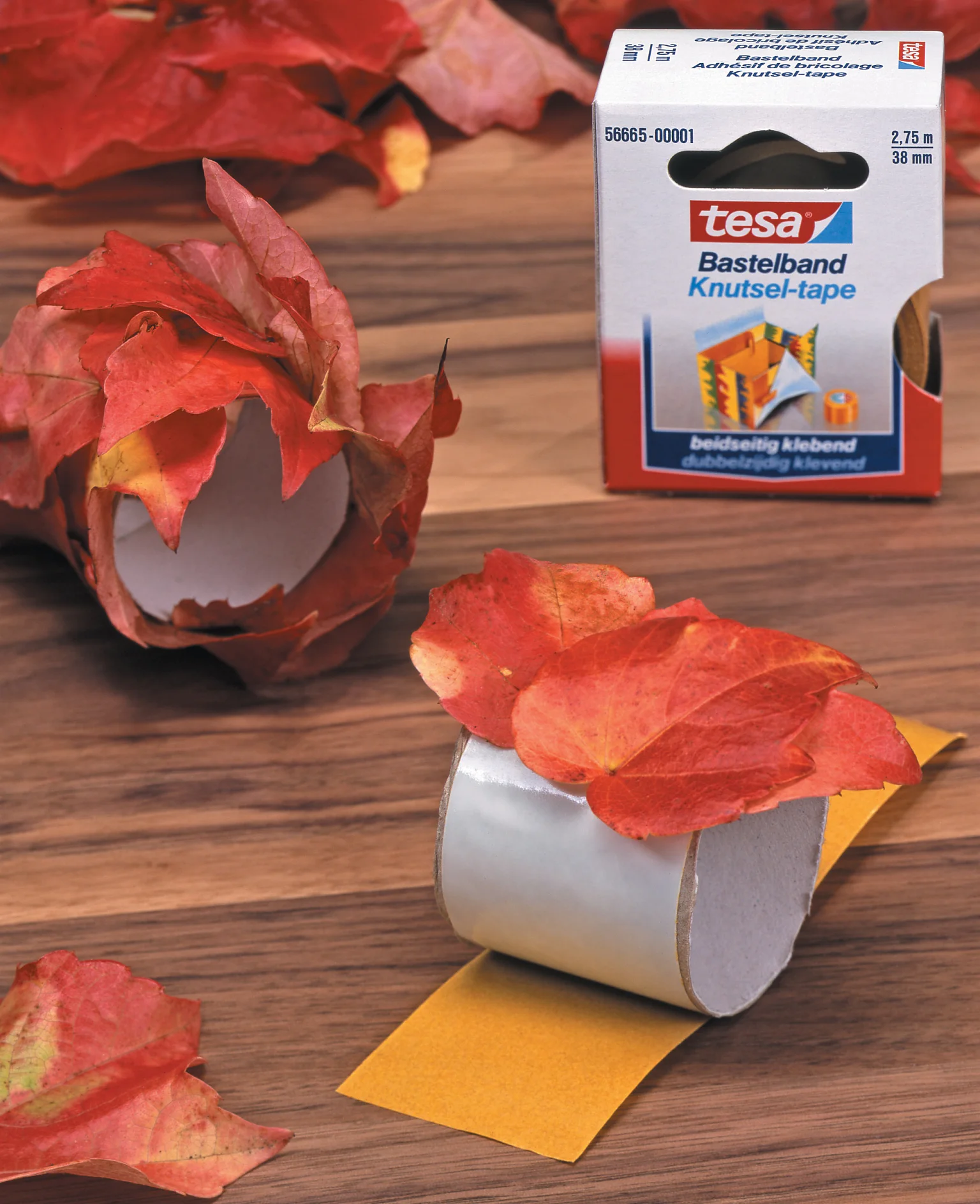 Een rol tesa tape dubbelzijdige kleefstof met een met bladeren bedekte rol ernaast en verschillende herfstbladeren verspreid op een houten oppervlak. De productverpakking is zichtbaar op de achtergrond. (This text has been generated by AI)