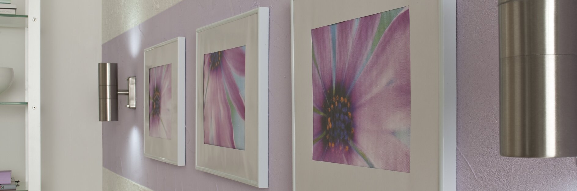 Drie ingelijste fotos van roze bloemen hangen aan een muur met een paarse horizontale streep, vastgezet met tesa tape. De muur is getextureerd en heeft twee metalen wandlampen. Een kast met glazen planken is gedeeltelijk zichtbaar aan de linkerkant. (This text has been generated by AI)