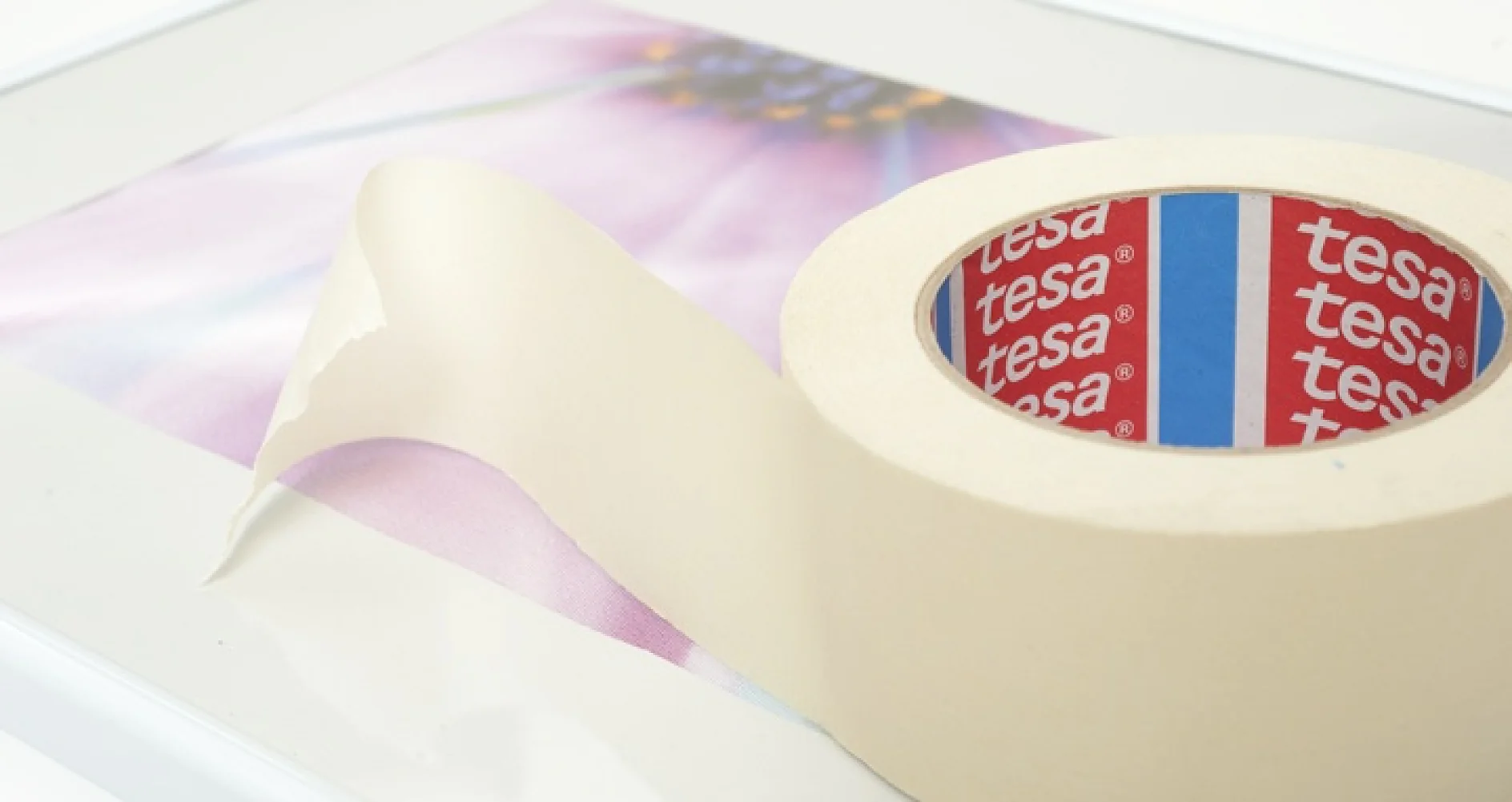 DIY Basics Een rol crèmekleurige tesa-tape wordt gedeeltelijk uitgerold en op een oppervlak gelegd met een wazige afbeelding van een kleurrijke bloem. (This text has been generated by AI)