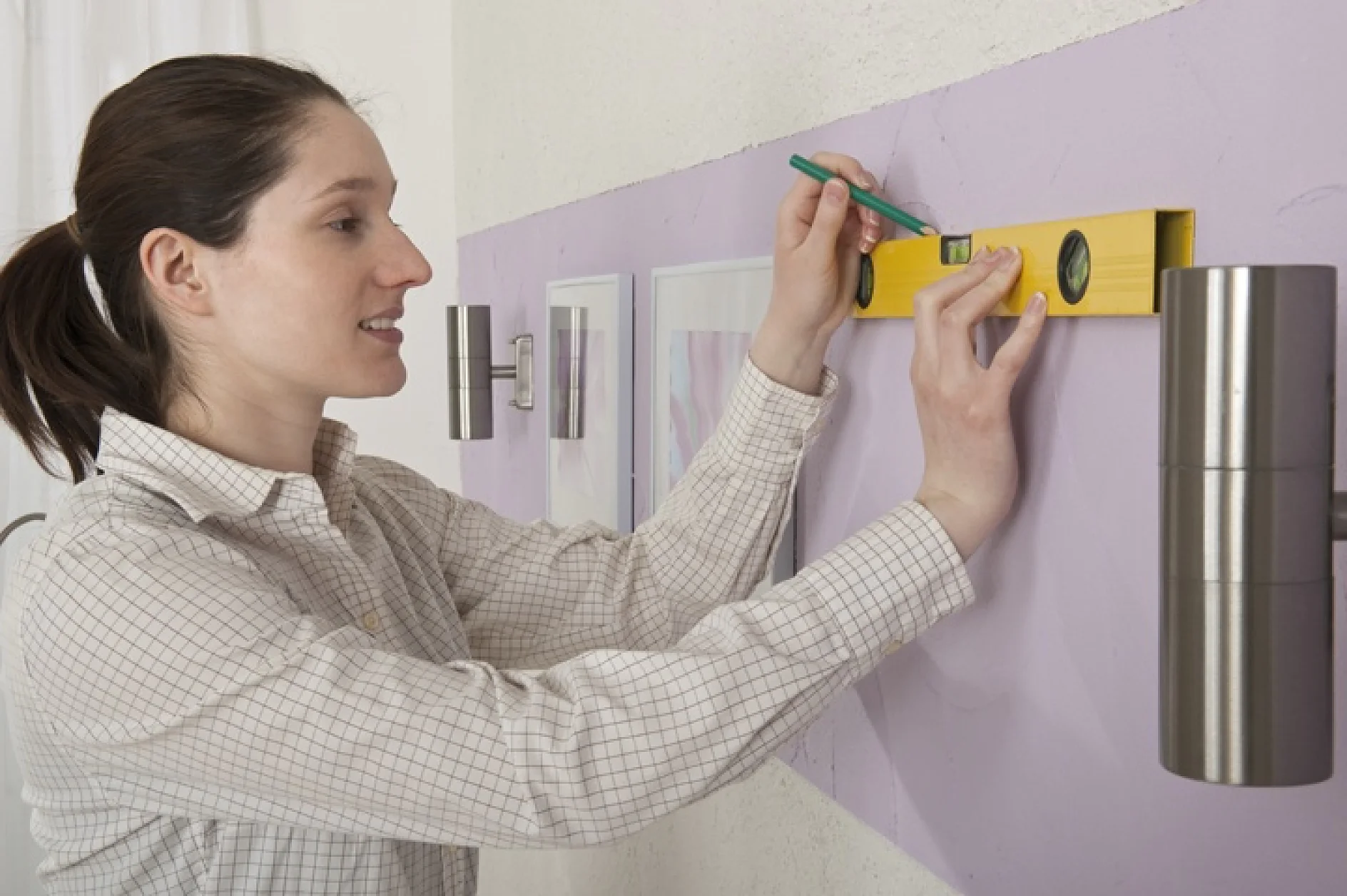 Een persoon die een geruit overhemd draagt, gebruikt een geel waterpasgereedschap op een paarse muur terwijl hij een ingelijste foto met tesa-tape aanpast. Aan beide kanten zijn twee aan de muur bevestigde lampen zichtbaar. (This text has been generated by AI)