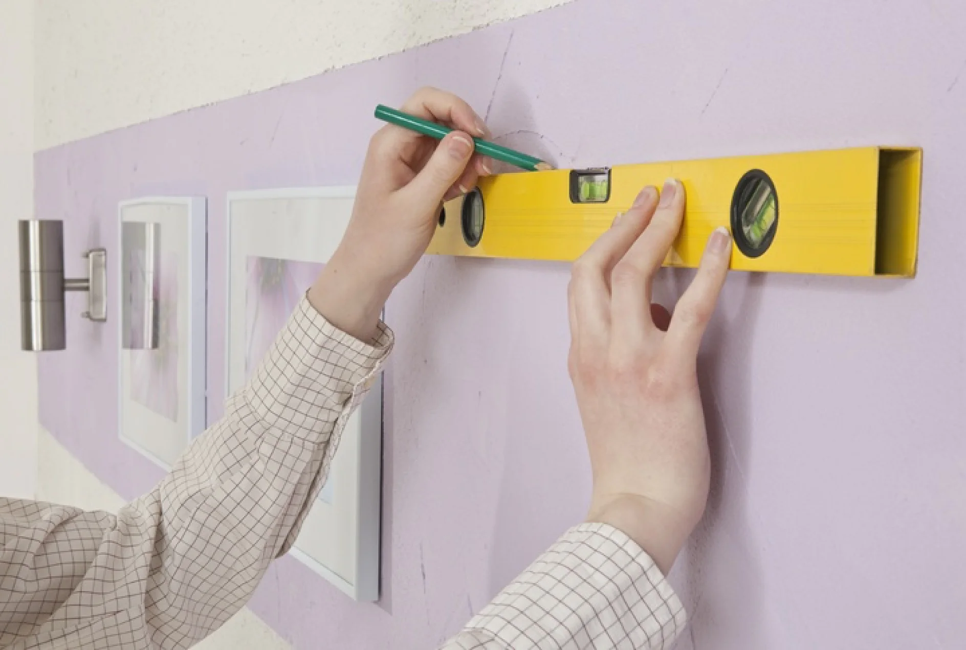 Een persoon in een geruit overhemd gebruikt een geel waterpasgereedschap om een lijn op een paarse muur te markeren met een groen potlood. Twee ingelijste fotos en een cilindervormige wandlamp zijn zichtbaar op de muur met tesa-tape. (This text has been generated by AI)