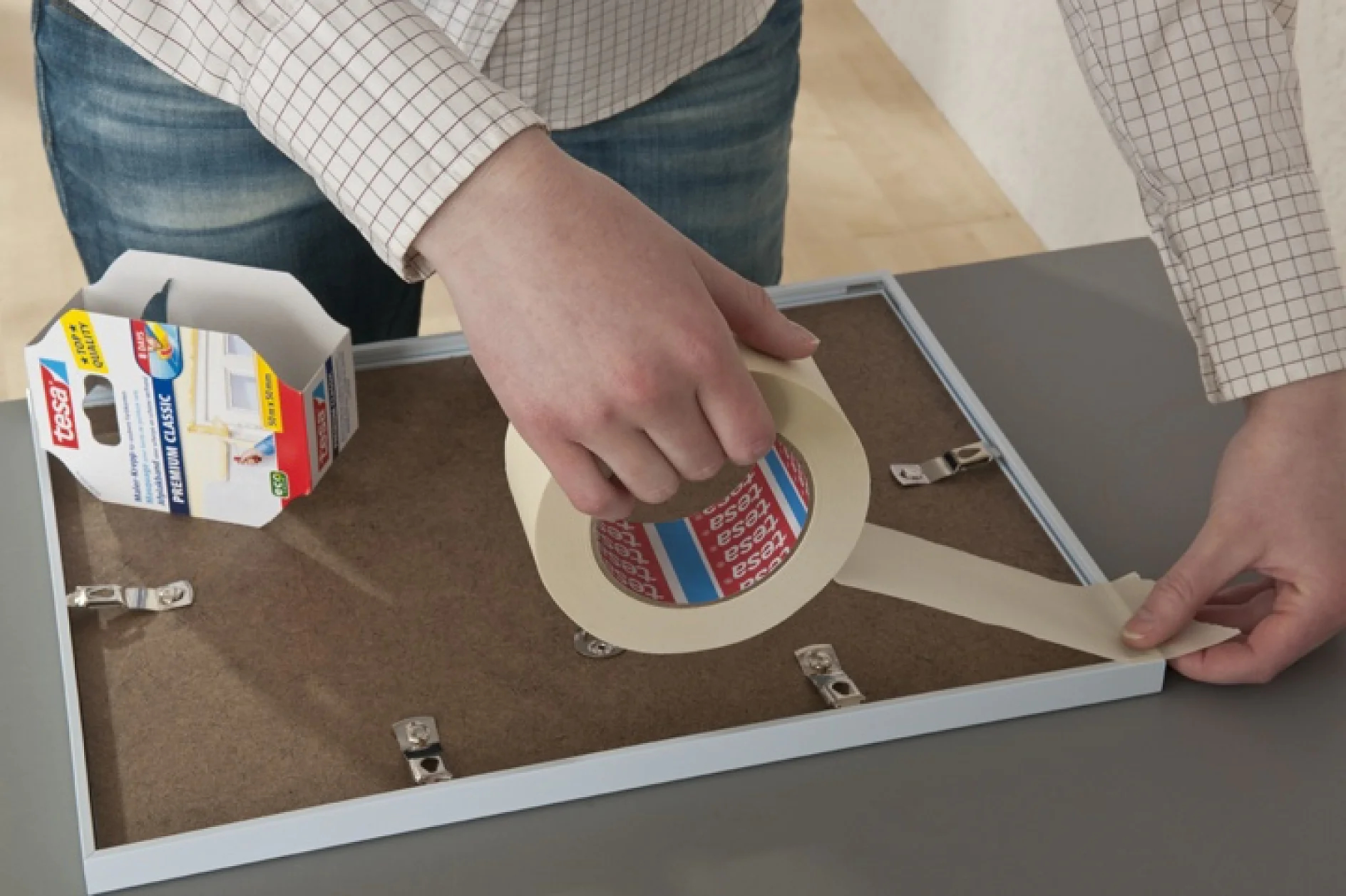 Een persoon brengt dubbelzijdige tesa tape aan op de achterkant van een fotolijst. De rol tesa tape zit in de linkerhand, terwijl de rechterhand de tesa tape op de rand van de lijst drukt. De doos voor de tesa tape staat op de tafel, gedeeltelijk open. (This text has been generated by AI)