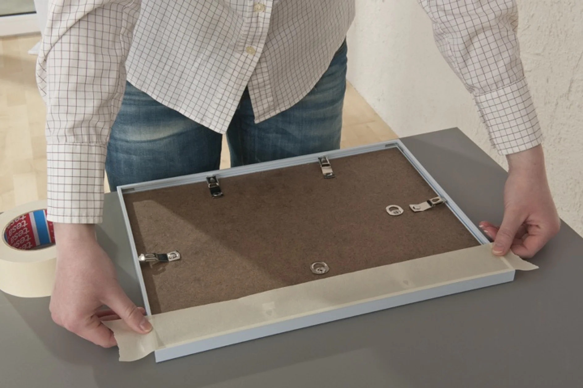 Een persoon bereidt een fotolijst voor om op te hangen door tesa tape langs de randen aan te brengen. De lijst staat op een grijze tafel en de persoon draagt een geruit overhemd en jeans. Een rol tesa tape is links zichtbaar. (This text has been generated by AI)
