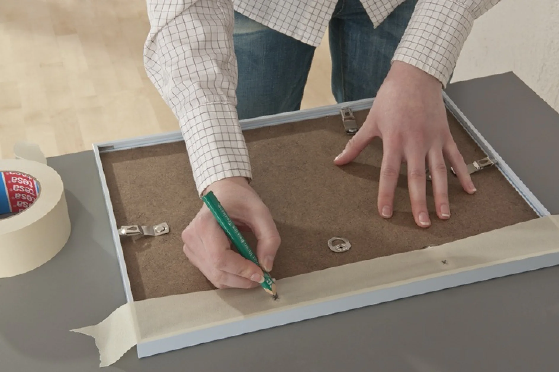 Een persoon markeert posities op de achterkant van een fotolijst met een potlood, op een tafel. Ze dragen een geruit overhemd en jeans. Een rol tesa tape en montagebeugels zijn in de buurt zichtbaar. (This text has been generated by AI)