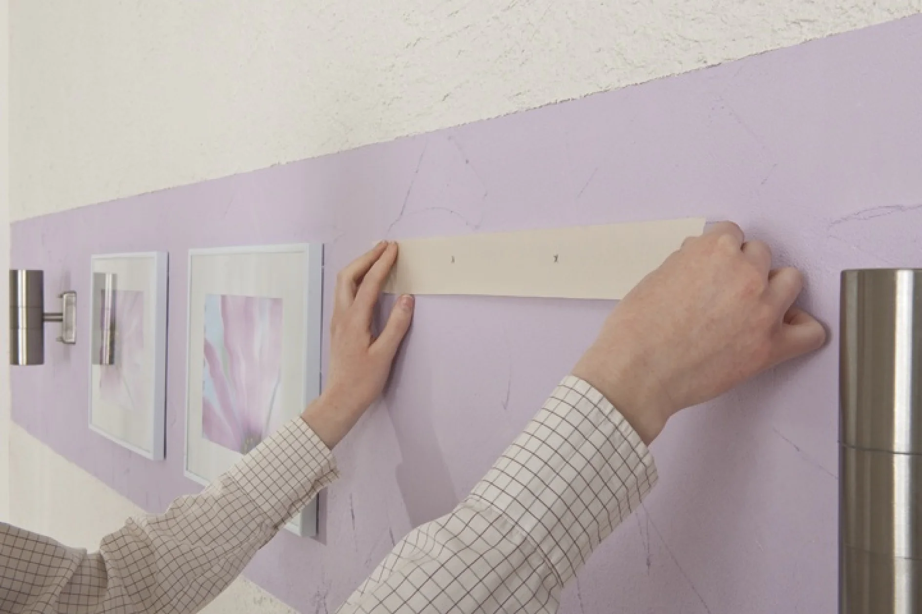 Iemand in een geruit overhemd hangt met tesa-tape een strook papier op een paarse muur, boven ingelijste fotos. (This text has been generated by AI)
