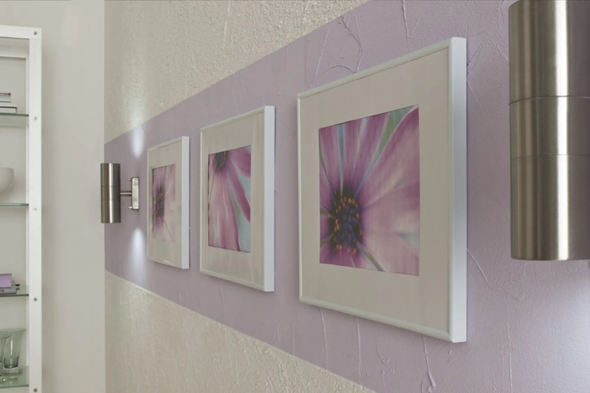 Drie ingelijste bloemenprints met roze en paarse bloemen hangen op een rij aan een getextureerde muur met een horizontale lavendelstreep. Twee moderne metalen wandlampen zijn aan beide kanten van de frames gemonteerd, stevig vastgezet met tesa-tape. (This text has been generated by AI)